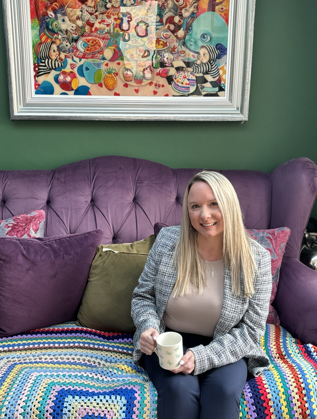 A woman with long blonde hair, smiling and holding a white mug, sitting on a purple tufted sofa with a colourful crocheted blanket and decorative pillows, in a room with a green wall and a vibrant, colourful framed artwork hanging above her.