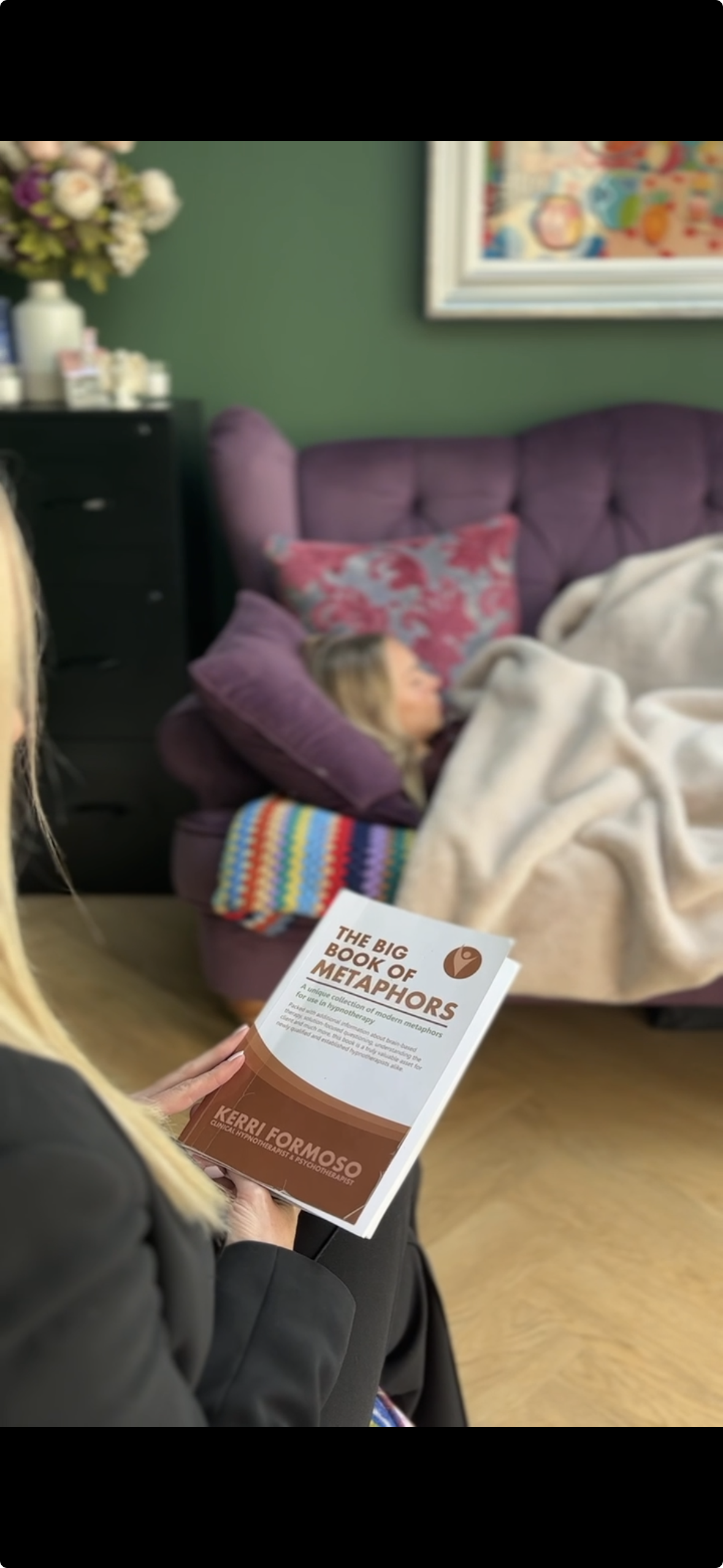 A woman holding a book titled 'The Big Book of Metaphors' sitting in a room with a woman lying on a purple couch, covered with a blanket, with colourful pillows behind her.