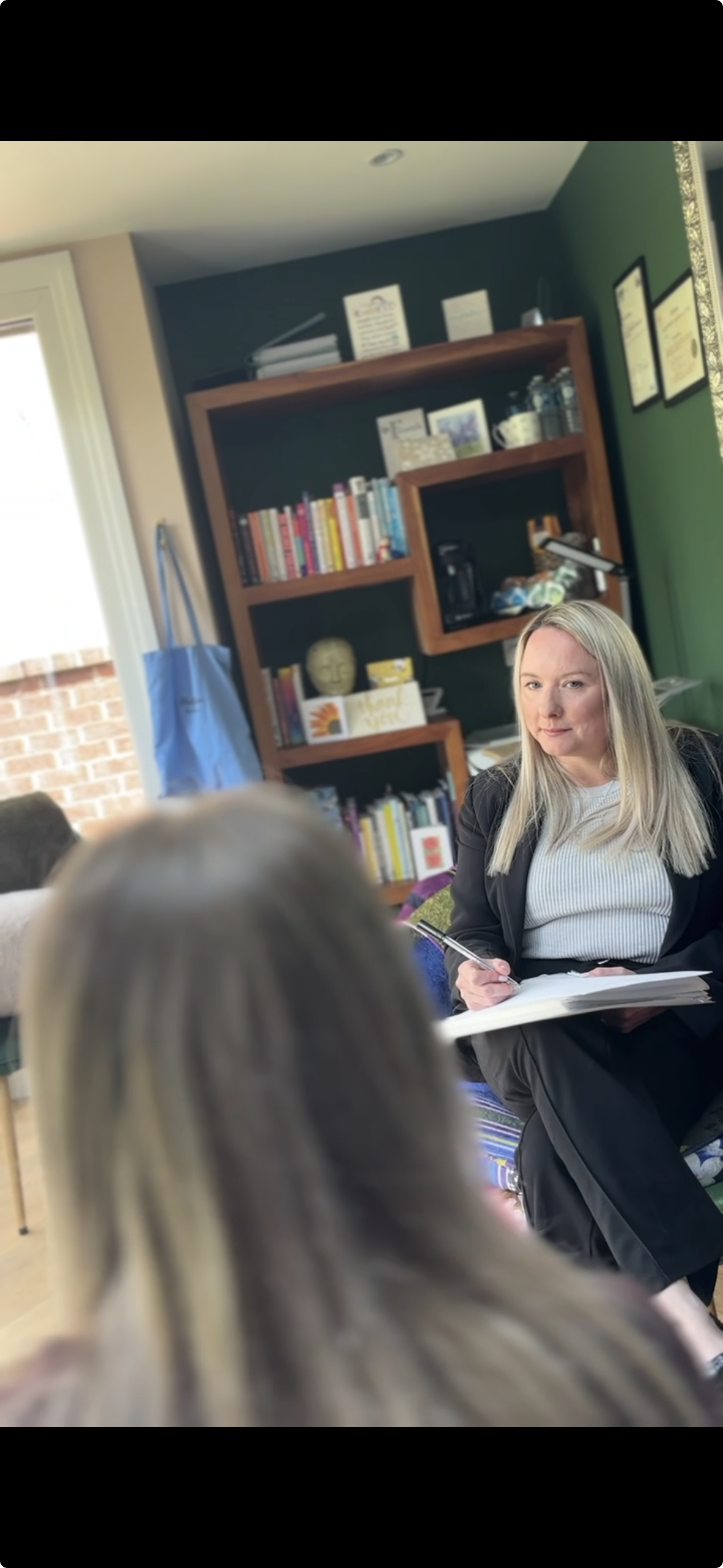 A woman sitting on a chair holding a pen and notebook, engaged in a conversation with a bookshelf filled with books and decorative items in the background.