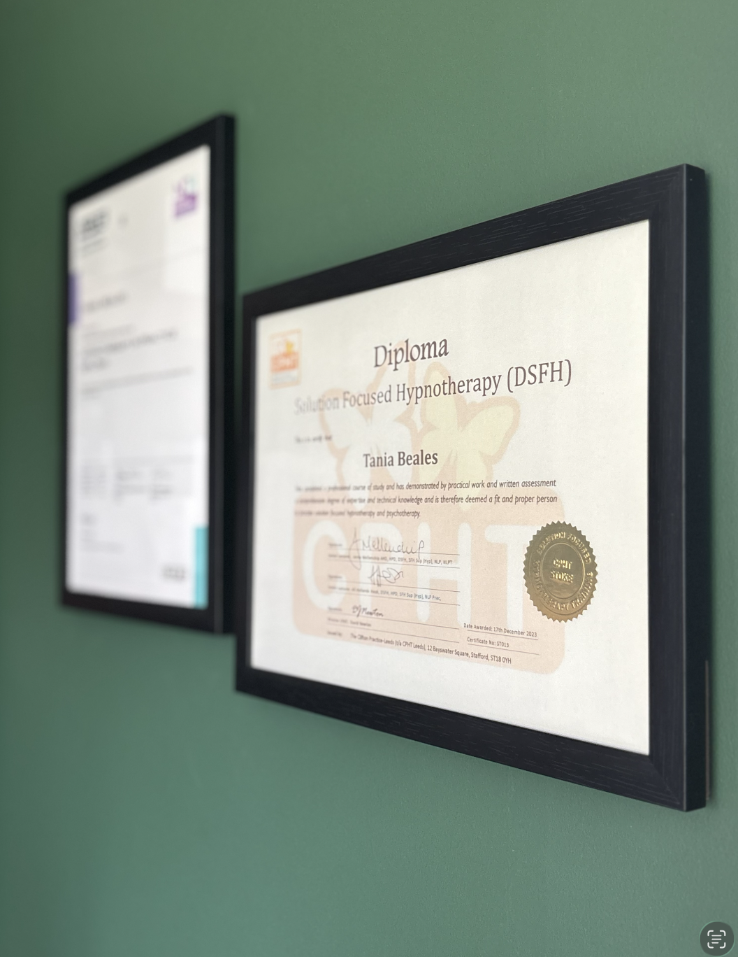 Close-up of a framed diploma from Solution Focused Hypnotherapy, awarded to Tania Beales, with a gold seal, hanging on a green wall, with another blurred framed certificate beside it.