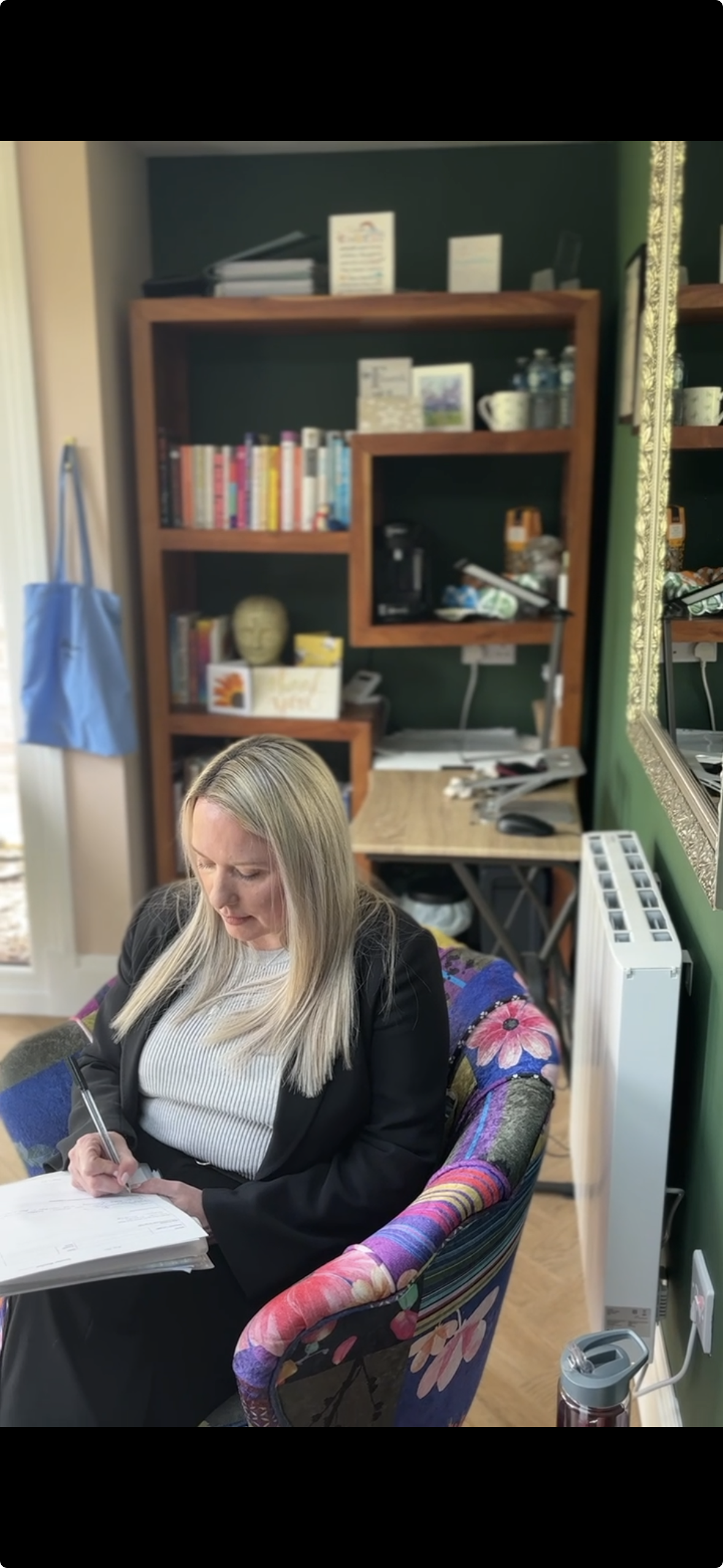 A woman with long blonde hair sitting in a colourful floral armchair, writing on papers with a pen, in an office space with a dark green wall, a wooden bookshelf in the background, a desk with assorted items, and a large mirror on the wall.