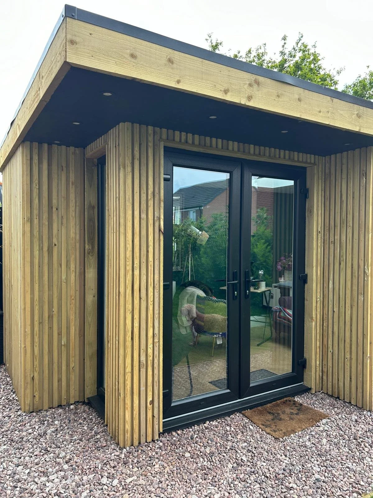 Small modern wooden garden room with glass double doors, situated on gravel, with a mat outside. Inside, a cosy workspace with a chair, table, and lamp, visible through the glass doors. Green trees and neighboring houses in the background.