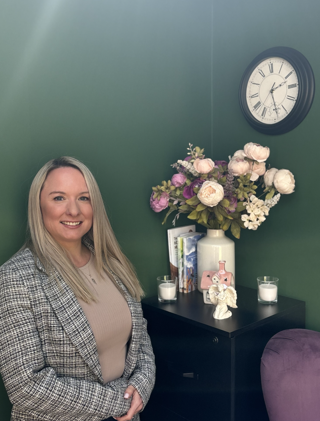 A woman with long blonde hair smiling, standing indoors next to a black cabinet with decorative items including a vase of pink and white flowers, candles, books, a pink angel figurine, and a white angel statue, against a green wall with a clock.