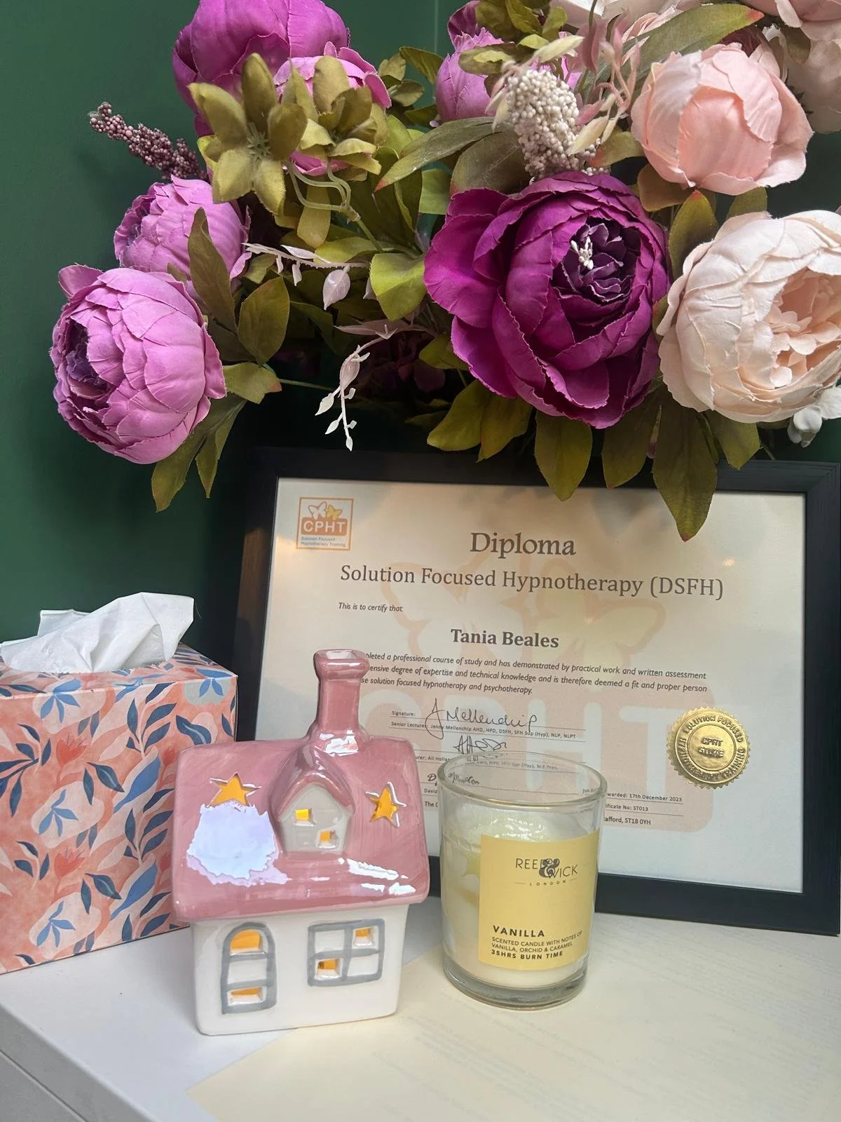 A bouquet of pink, purple, and cream artificial flowers next to a framed diploma for Solution Focused Hypnotherapy, a pink ceramic house ornament, a box of tissue, a yellow vanilla scented candle, and a glass of water on a white table.
