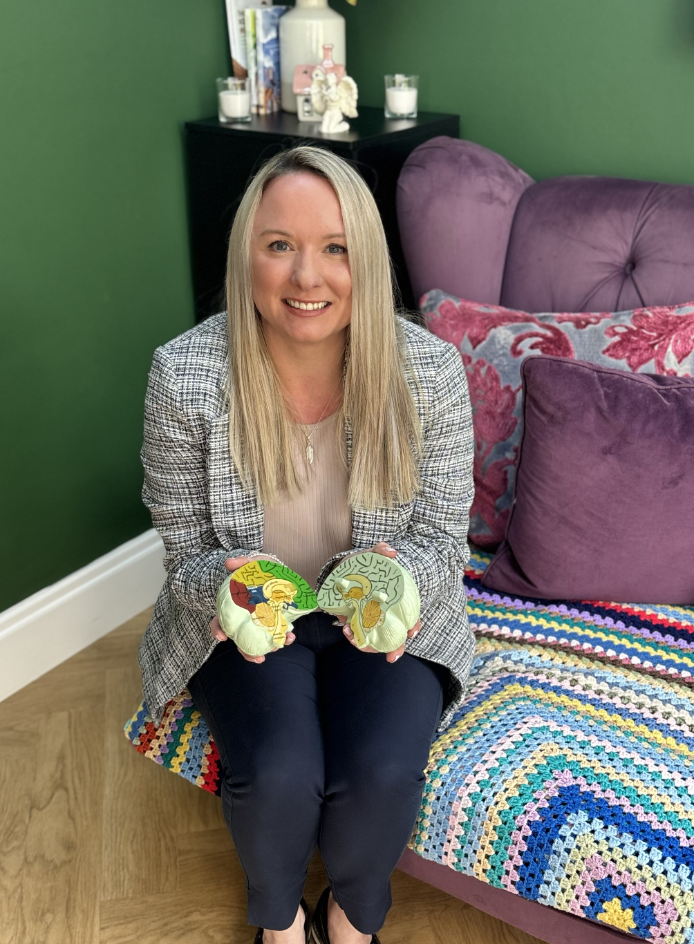 A woman sitting on a colourful crocheted blanket on a pink couch, holding two paper cutouts of human brain diagrams. She has long blonde hair, is smiling, and is dressed in a light-colored top with a checkered blazer and dark pants.