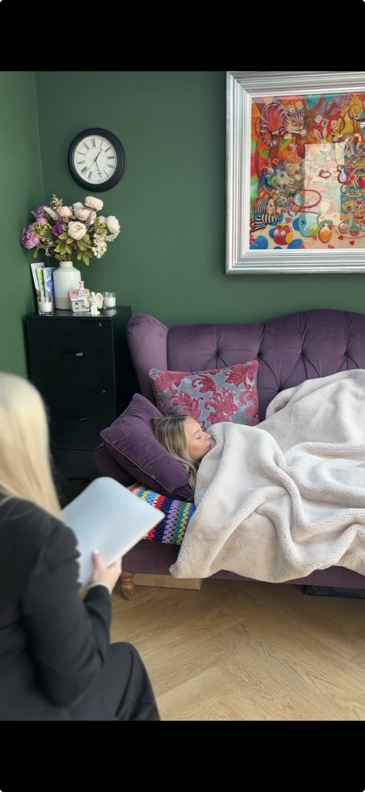 A young girl with blonde hair sleeping on a purple couch with a pink and red decorative pillow, covered with a beige blanket, in a green-walled room. A woman in a dark jacket is sitting on a chair nearby, holding a tablet. There is a black cabinet with a large flower arrangement, candles, and books, and a colorful framed artwork on the wall.