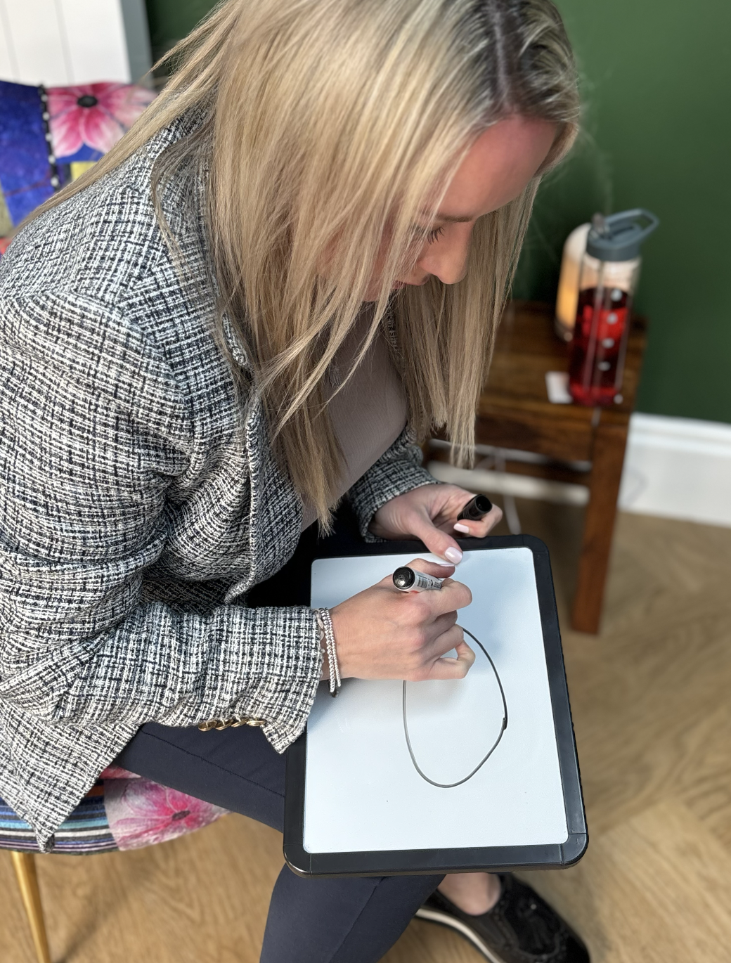 A woman with blonde hair, wearing a patterned blazer, is sitting on a chair, drawing a circle on a whiteboard with a marker.