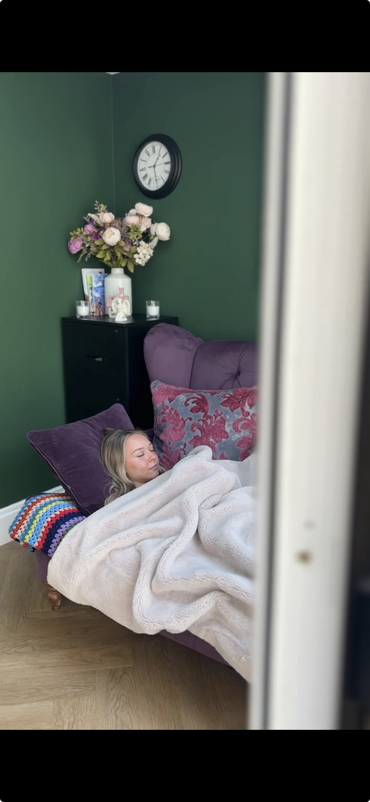 Young woman sleeping on a purple sofa. The sofa has two large pillows with pink and grey patterns. Behind her is a green wall with a black clock and a black nightstand with a large white vase of flowers, candles, and decorative items.