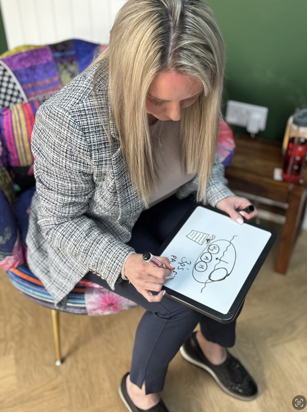 A woman with blonde hair, wearing a patterned blazer and black trainers, is sitting on a colourful chair and writing on a digital tablet with a black marker. The tablet displays a diagram or drawing.