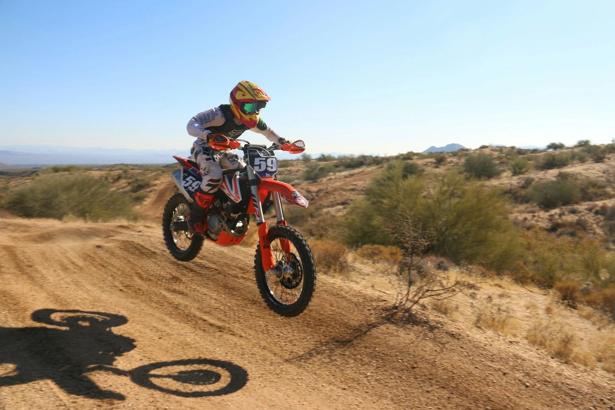 A person riding an orange dirt bike on a dirt track in a desert landscape with mountains in the background, under a clear blue sky.