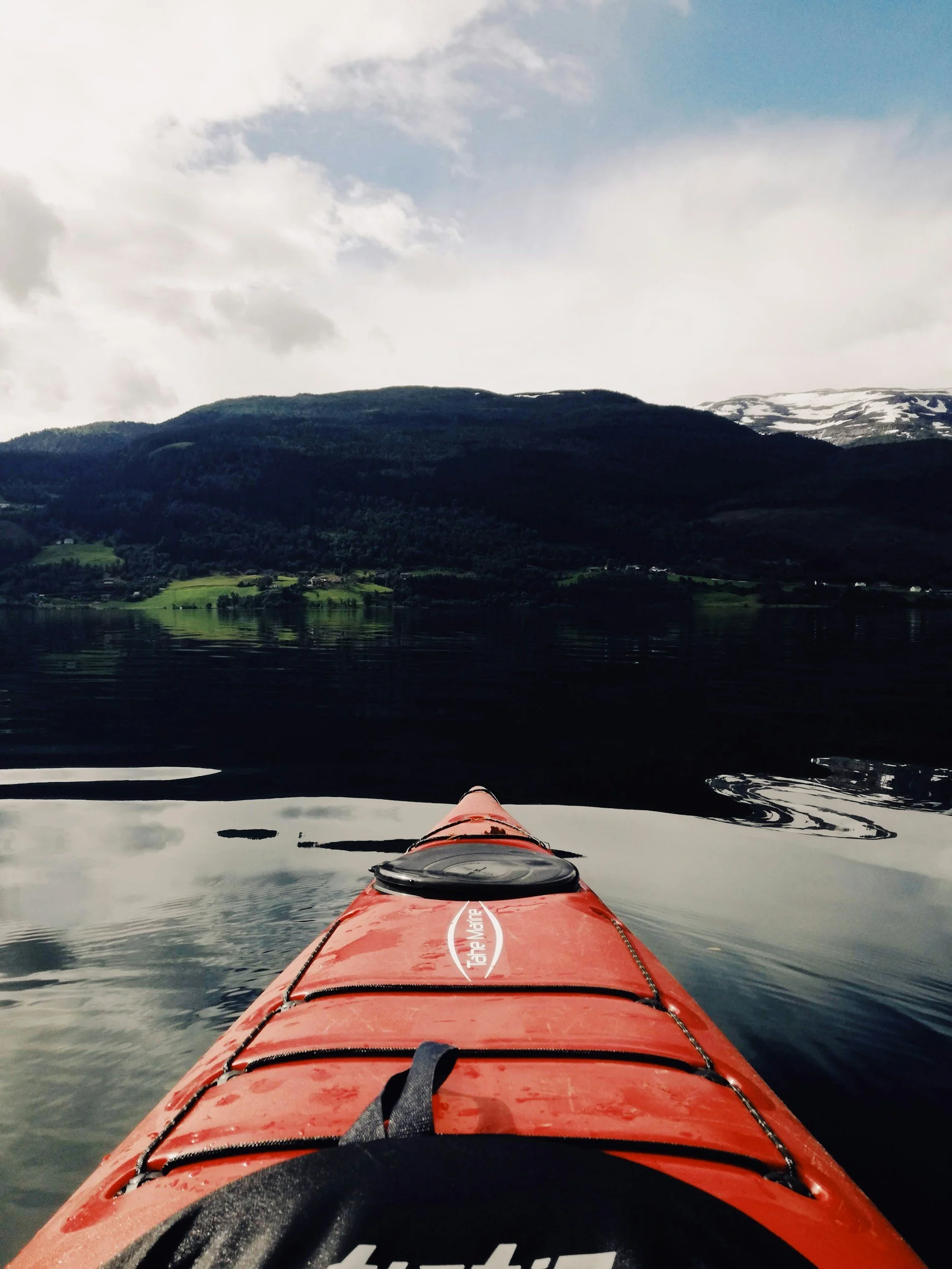 View from the front of a red kayak on calm water, with a distant green mountainside, partly cloudy sky, and snow patches on a mountain in the background.