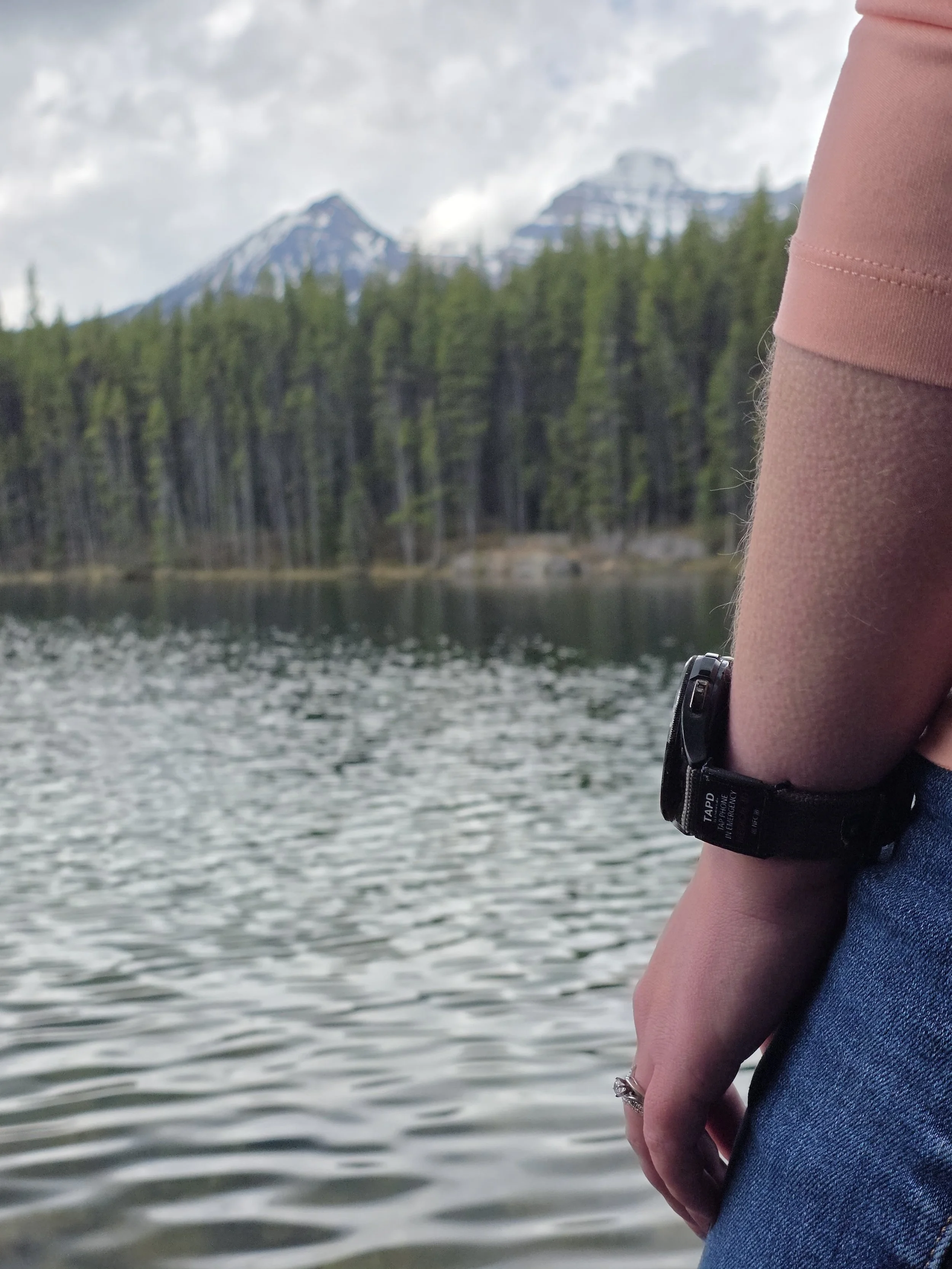 A close-up view of a person's arm with a wristwatch, wearing a peach-colored short sleeve shirt, next to a body of water with mountains and dense forest in the background.