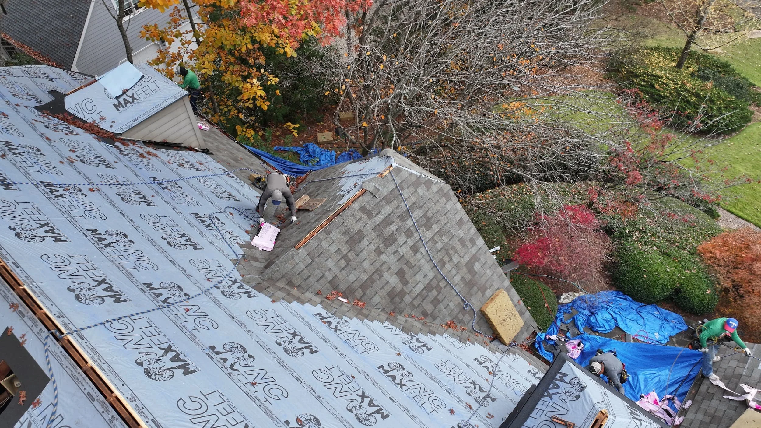 People installing shingles on a roof during a construction project, with autumn trees and a green yard in the background.