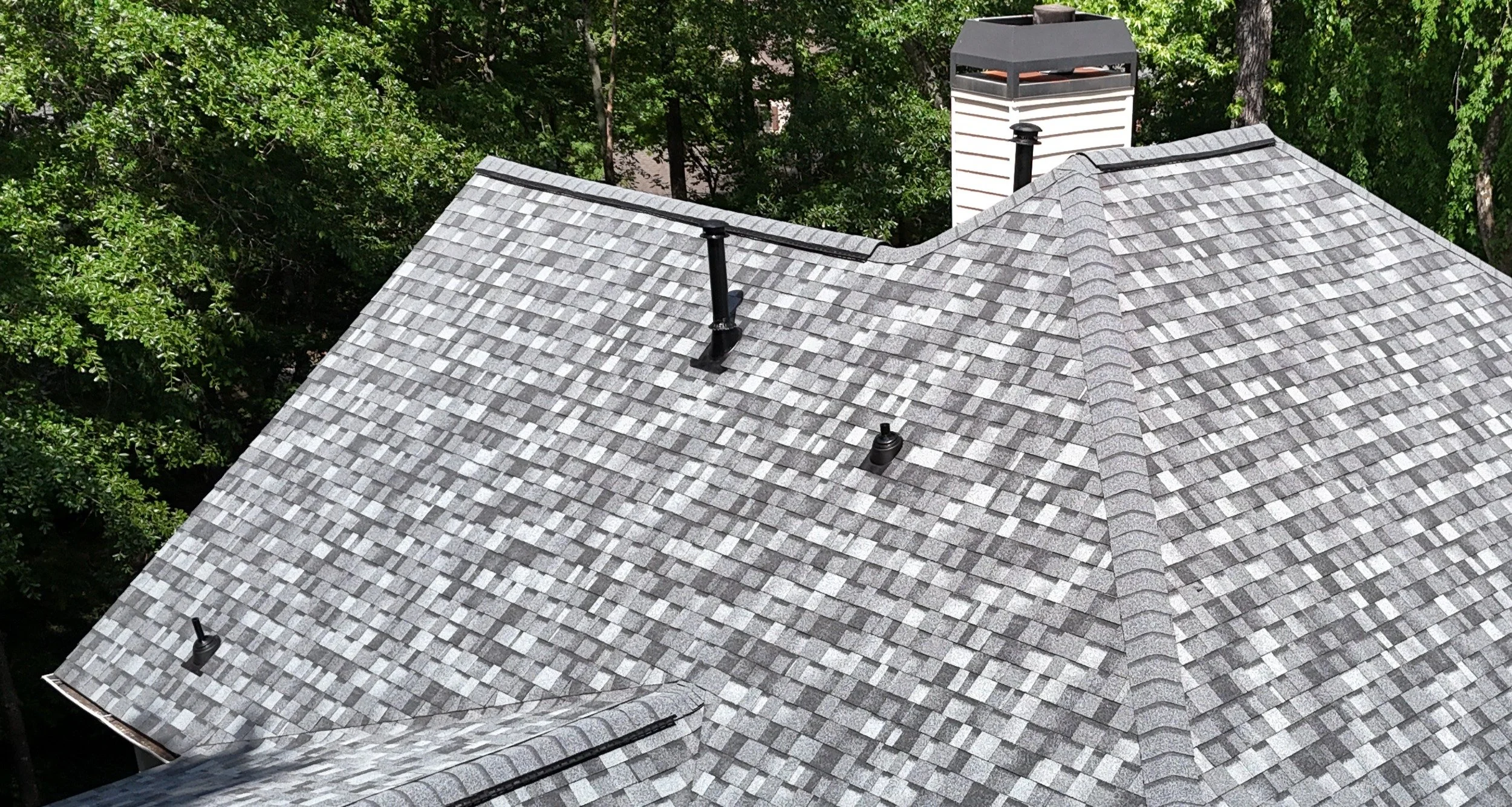 A rooftop with gray asphalt shingles, a chimney, and weather vanes, surrounded by green trees.