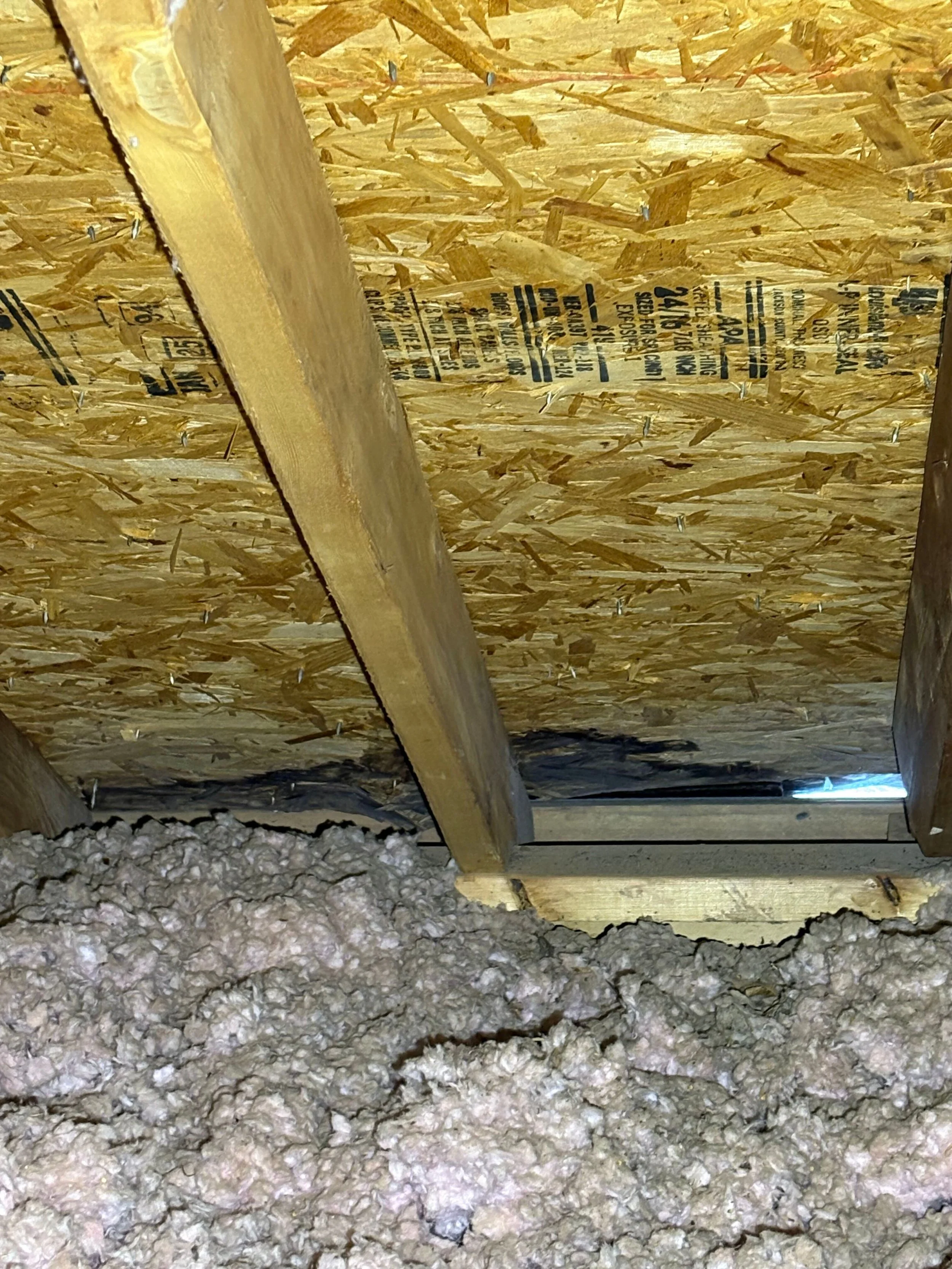 View of ceiling in attic or crawl space showing plywood sheathing, wooden beams, construction staples, insulation on the floor, and a small opening allowing natural light.