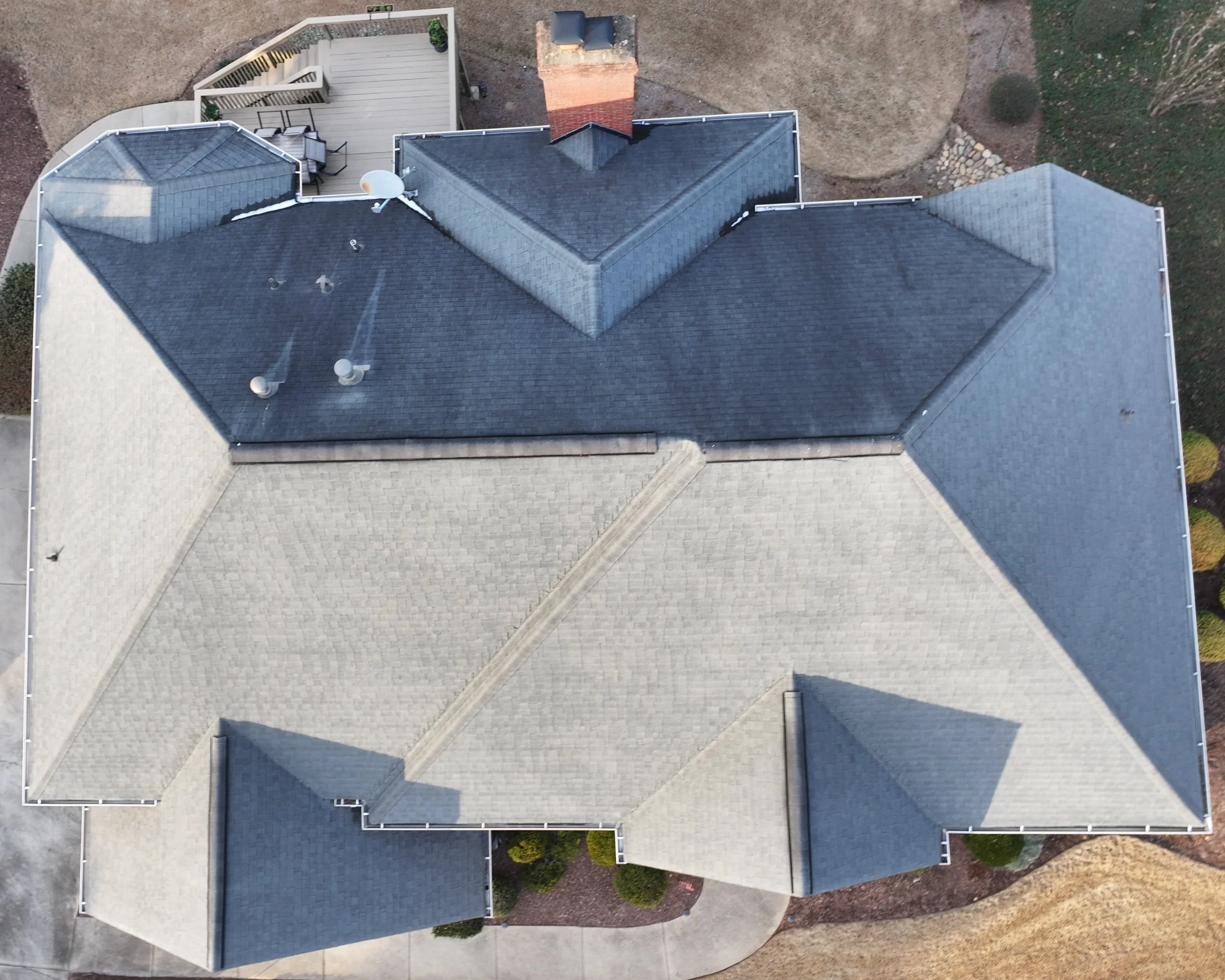 An aerial view of a house roof covered in dark shingles with a front porch and a brick chimney, surrounded by a yard with grass and patches of dirt.