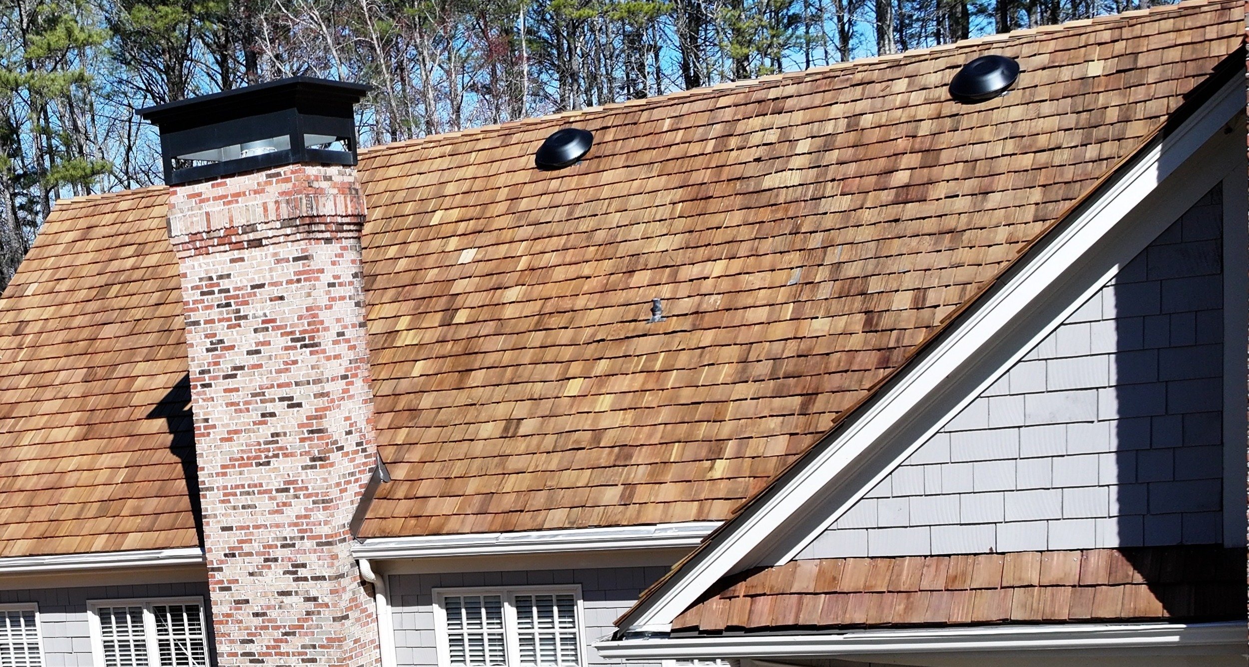 Close-up of a house roof with new tan shingles, a brick chimney, and a gable with white siding, surrounded by trees and a blue sky.