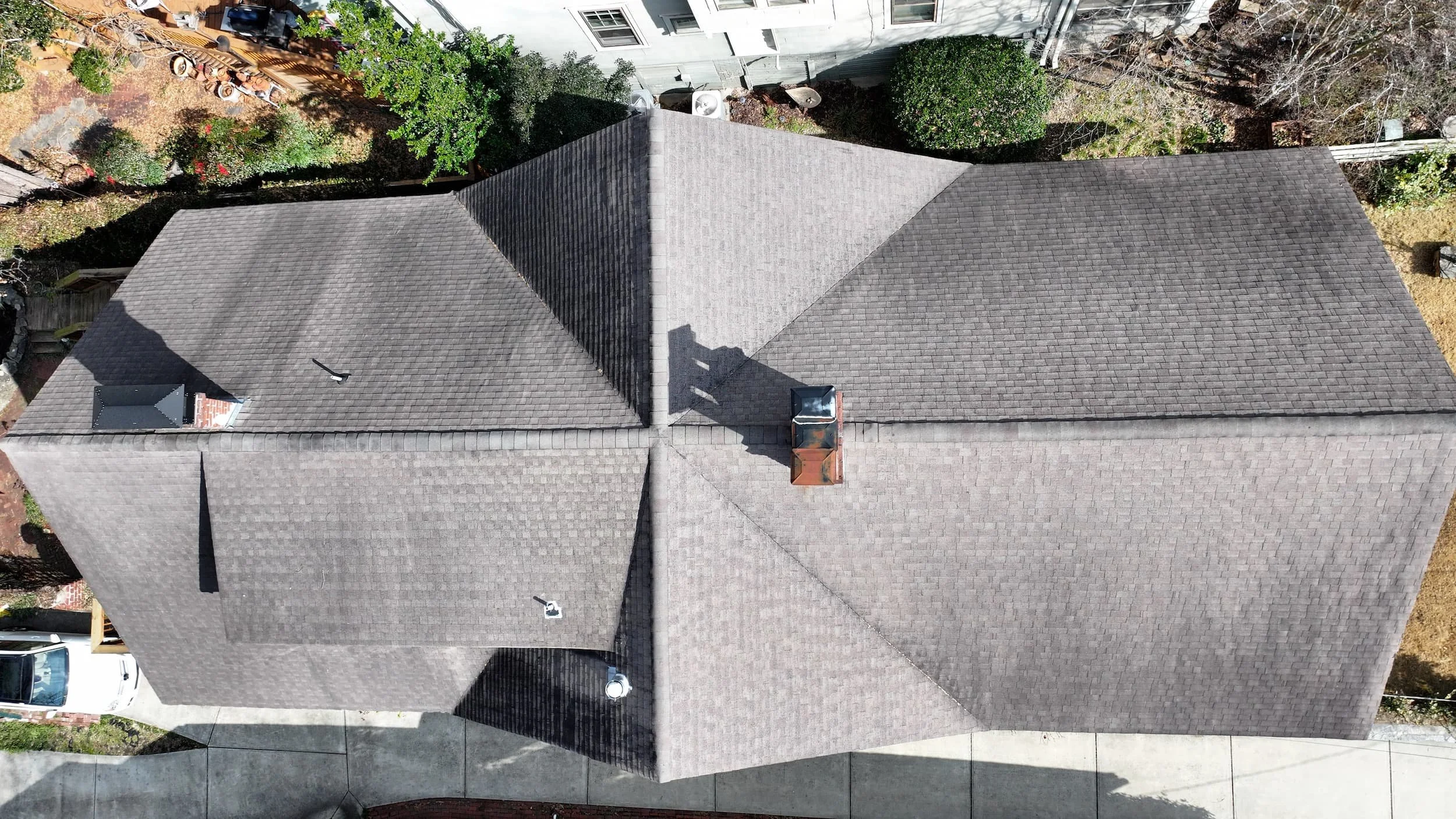An aerial view of a house's roof with multiple sections covered in gray shingles, with a chimney on one side and trees and neighboring buildings surrounding it.