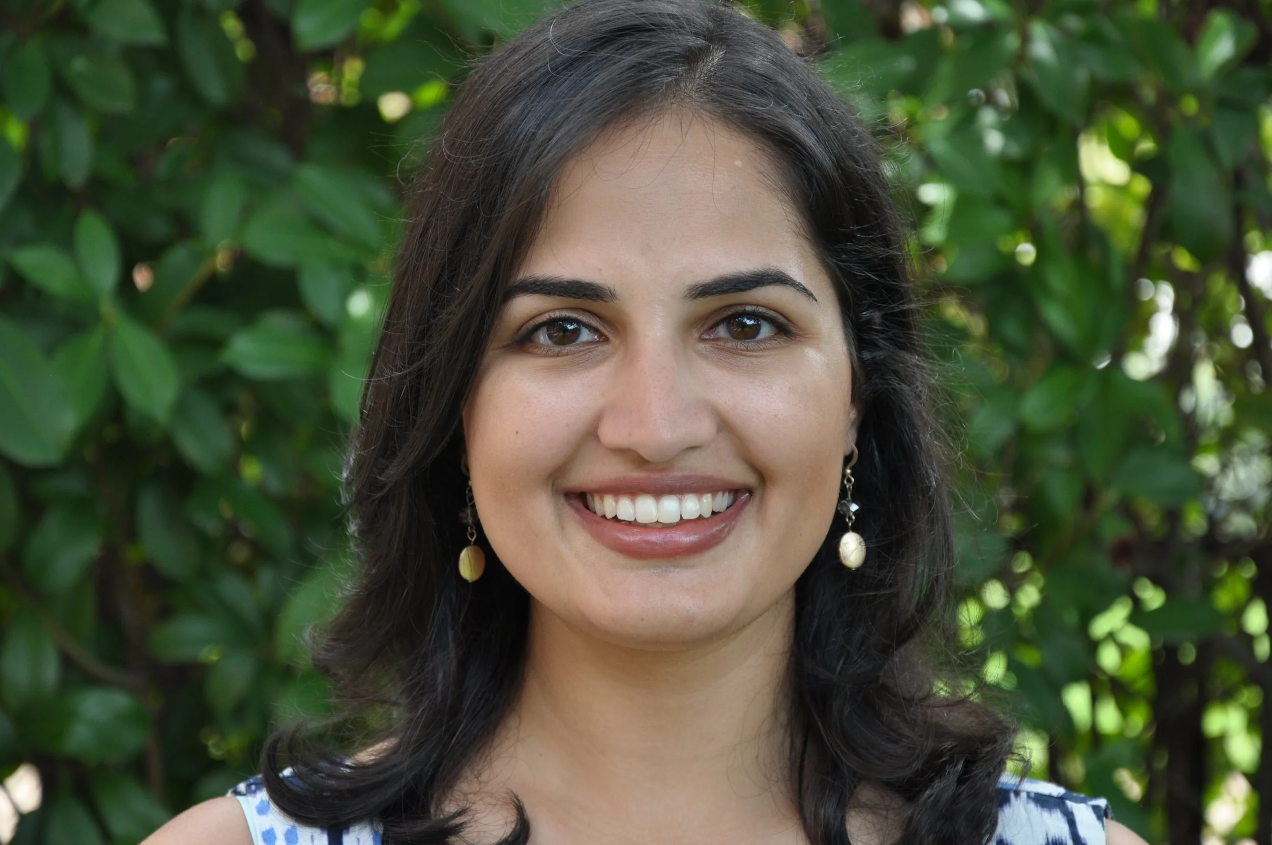 A woman with long dark hair and earrings smiling outdoors against a background of green foliage.