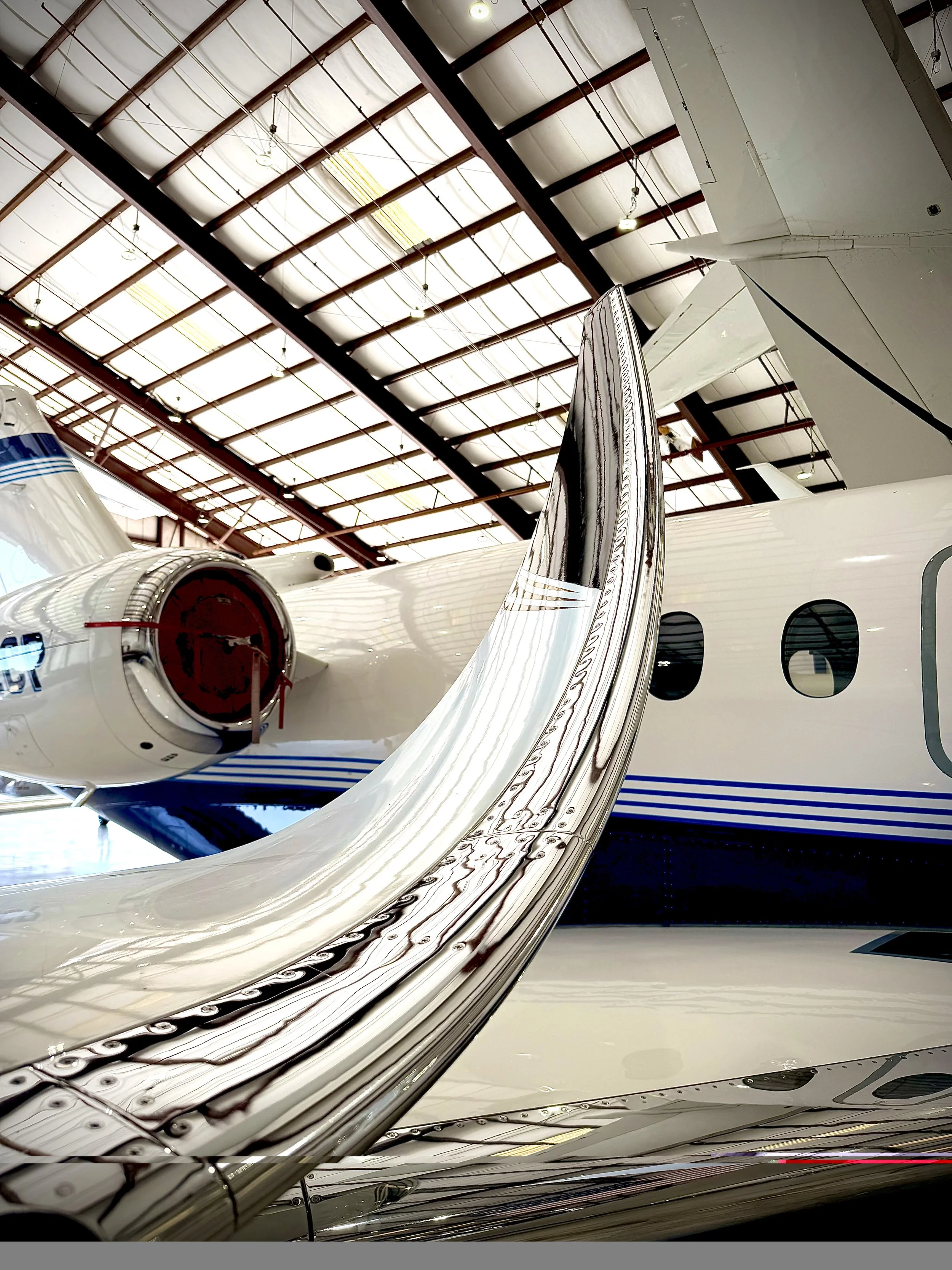 Inside an aircraft hangar, a white private jet with a shiny, reflective surface is parked. The image captures part of the aircraft's wing and engine, with the hangar's high ceiling and lighting visible above.