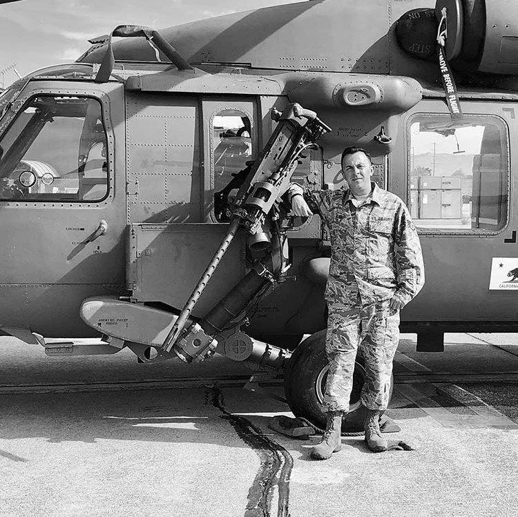 A soldier in camouflage uniform standing next to a military helicopter with a rocket launcher mounted underneath. The soldier is leaning on the helicopter's door frame with one hand, and the helicopter is parked on a concrete surface.