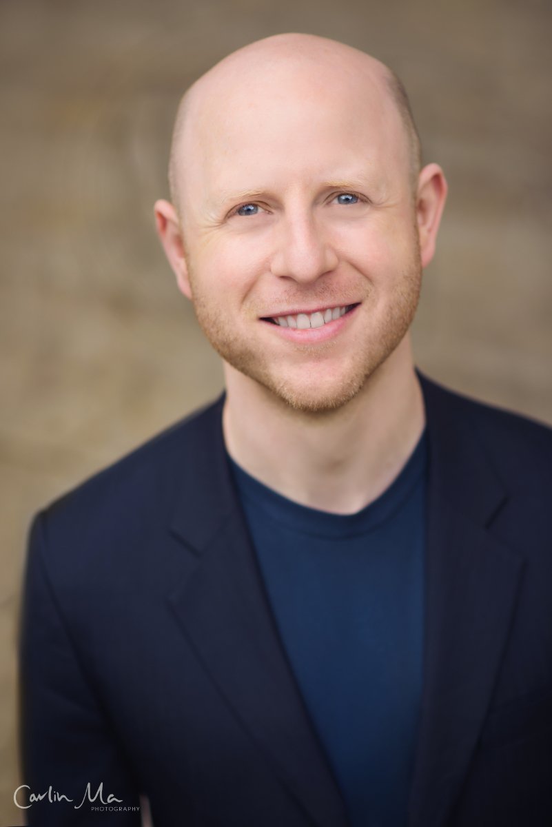 A professional headshot of a smiling man with a bald head, blue eyes, and light facial hair, wearing a dark blazer and navy shirt, against a blurry neutral background.