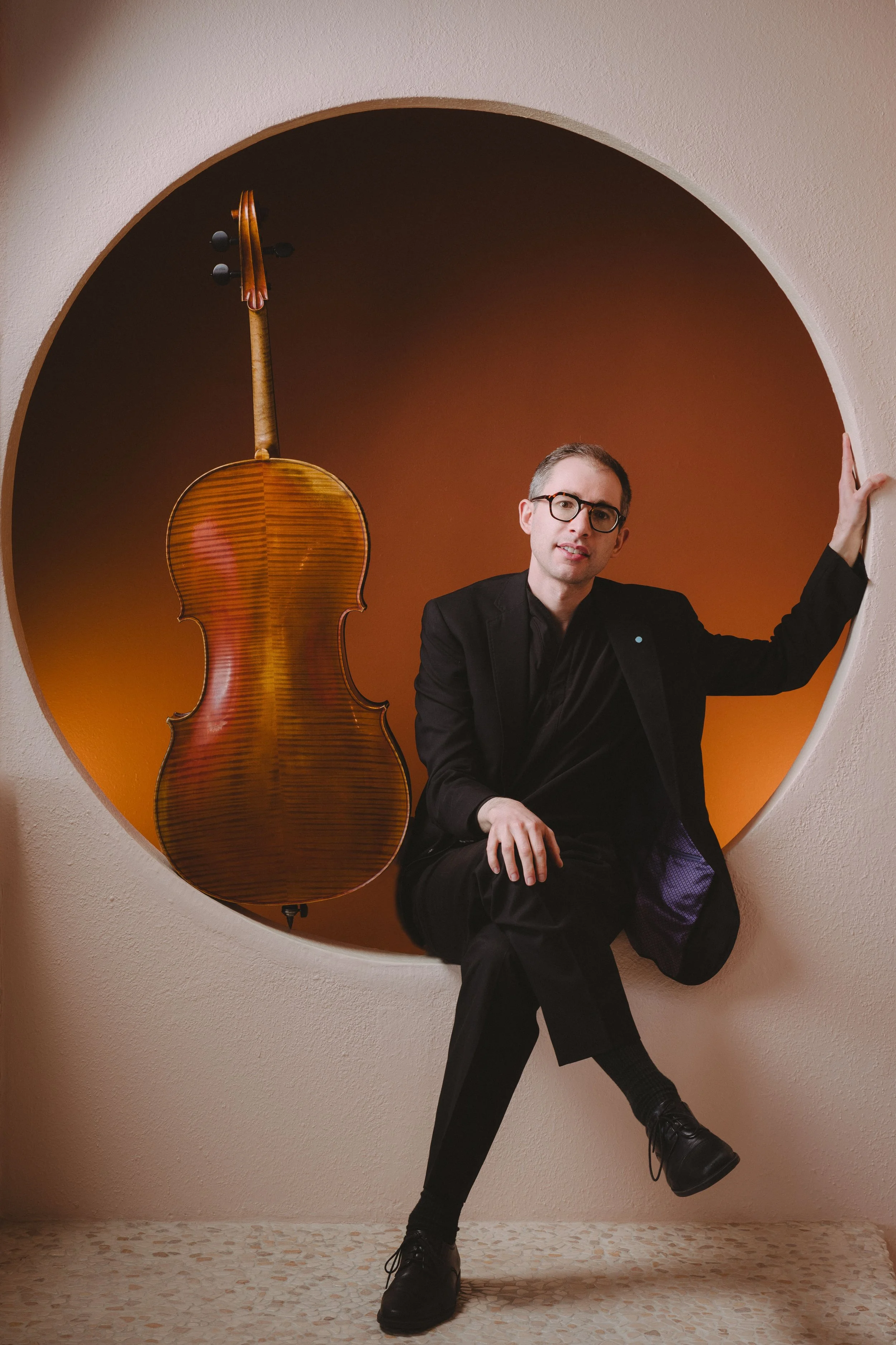 A man in a black suit and glasses sitting on a ledge with a cello hanging on the wall behind him, inside a circular cutout of a wall.
