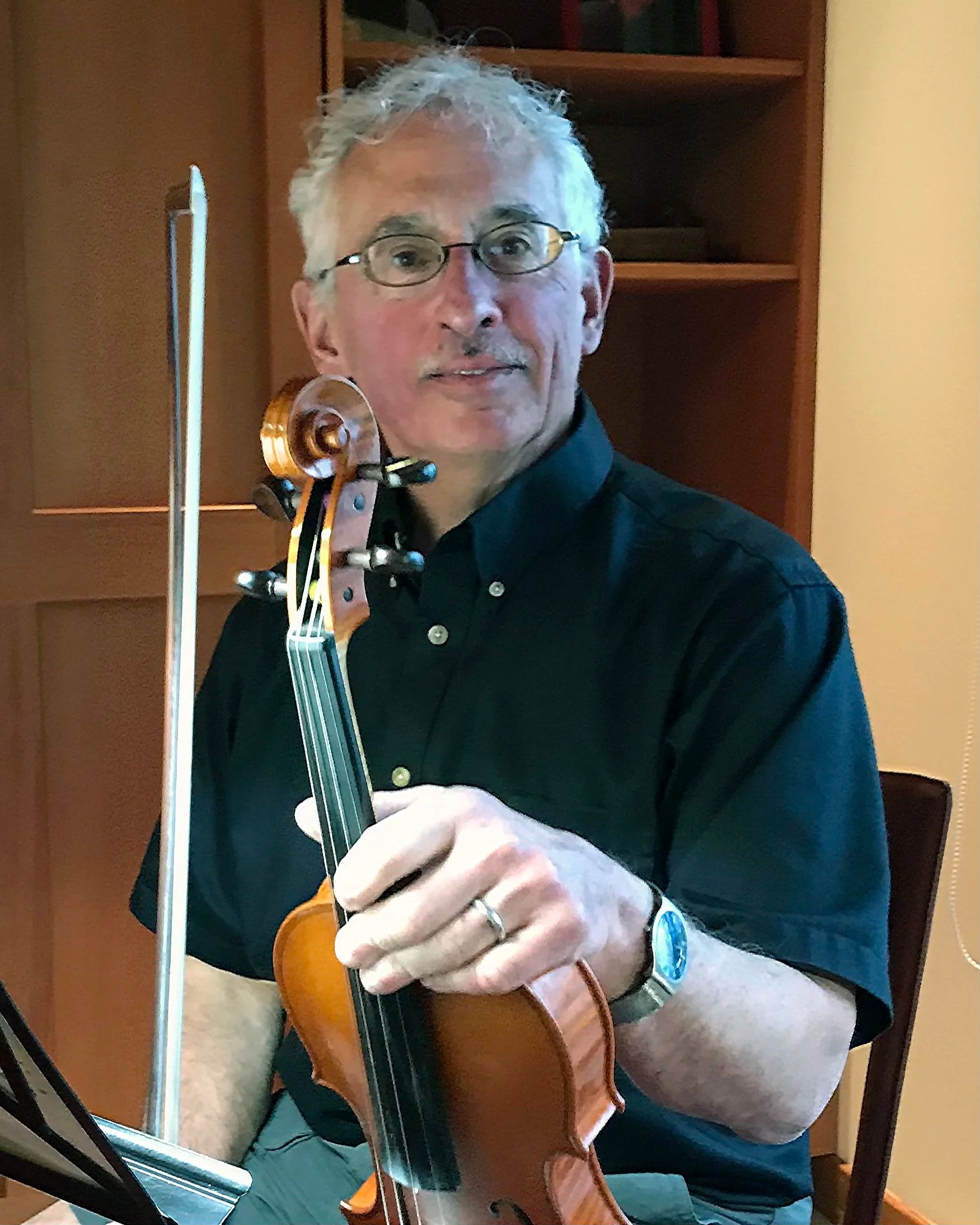 A man with glasses playing a violin, with a wooden bookshelf in the background.