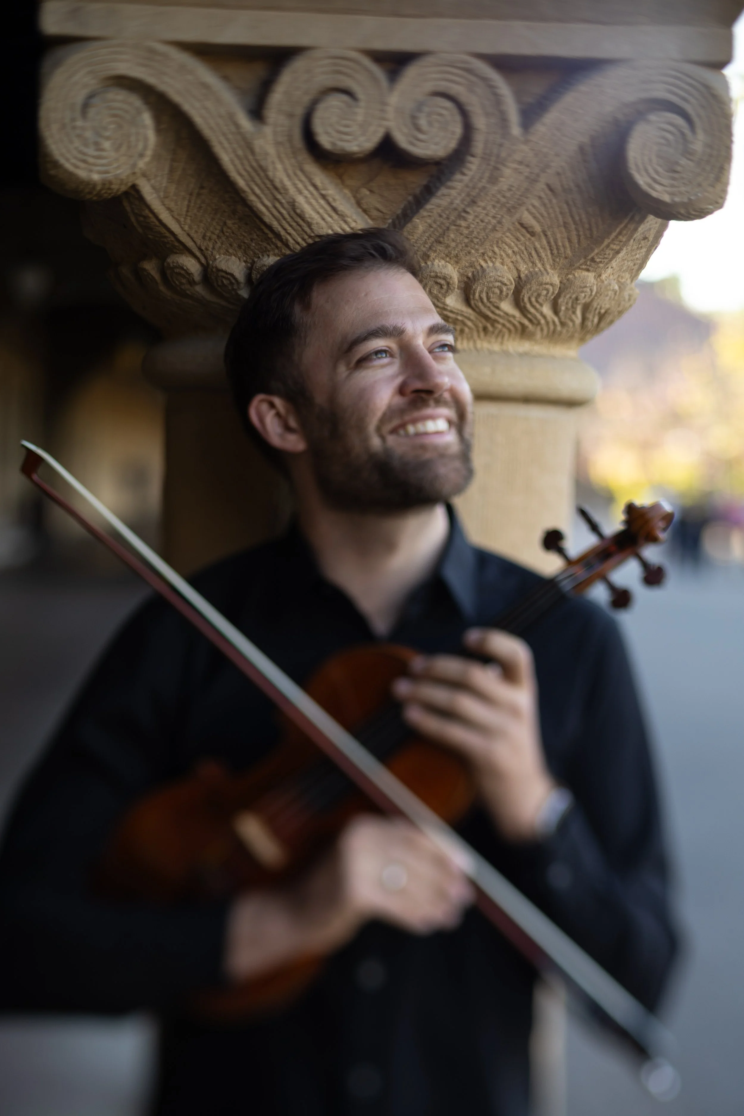 A man with short brown hair and a beard smiling, holding a violin and bow, standing under an ornate stone archway.