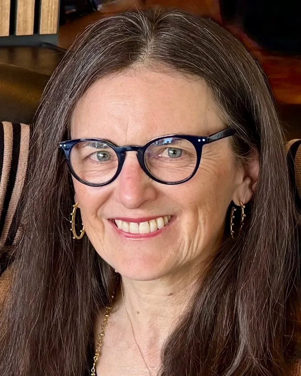 A woman with brown hair, wearing glasses, gold hoop earrings, and a gold necklace, smiling in an indoor setting.