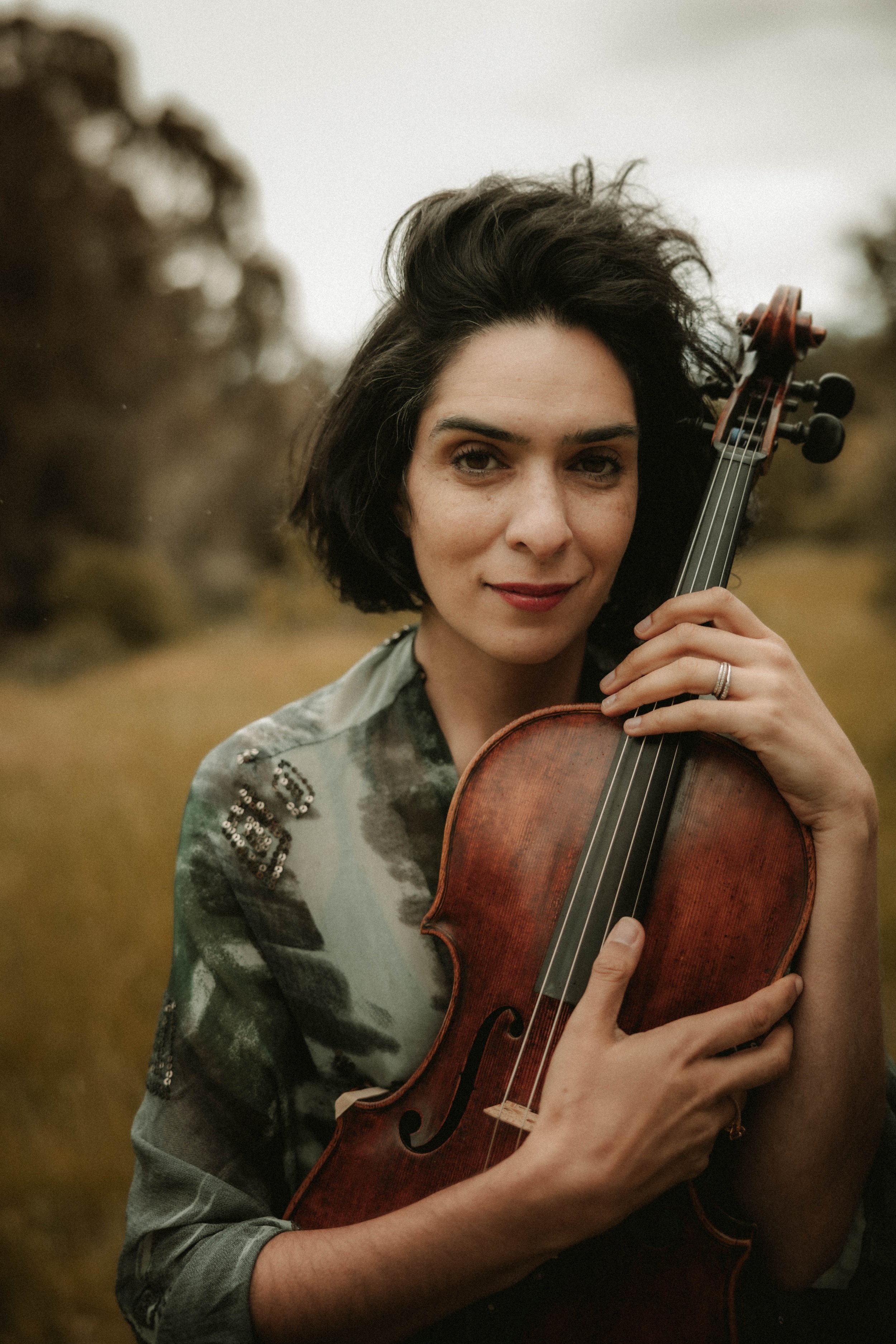 A woman with dark, curly hair playing a violin outdoors in a natural setting with blurred trees in the background.