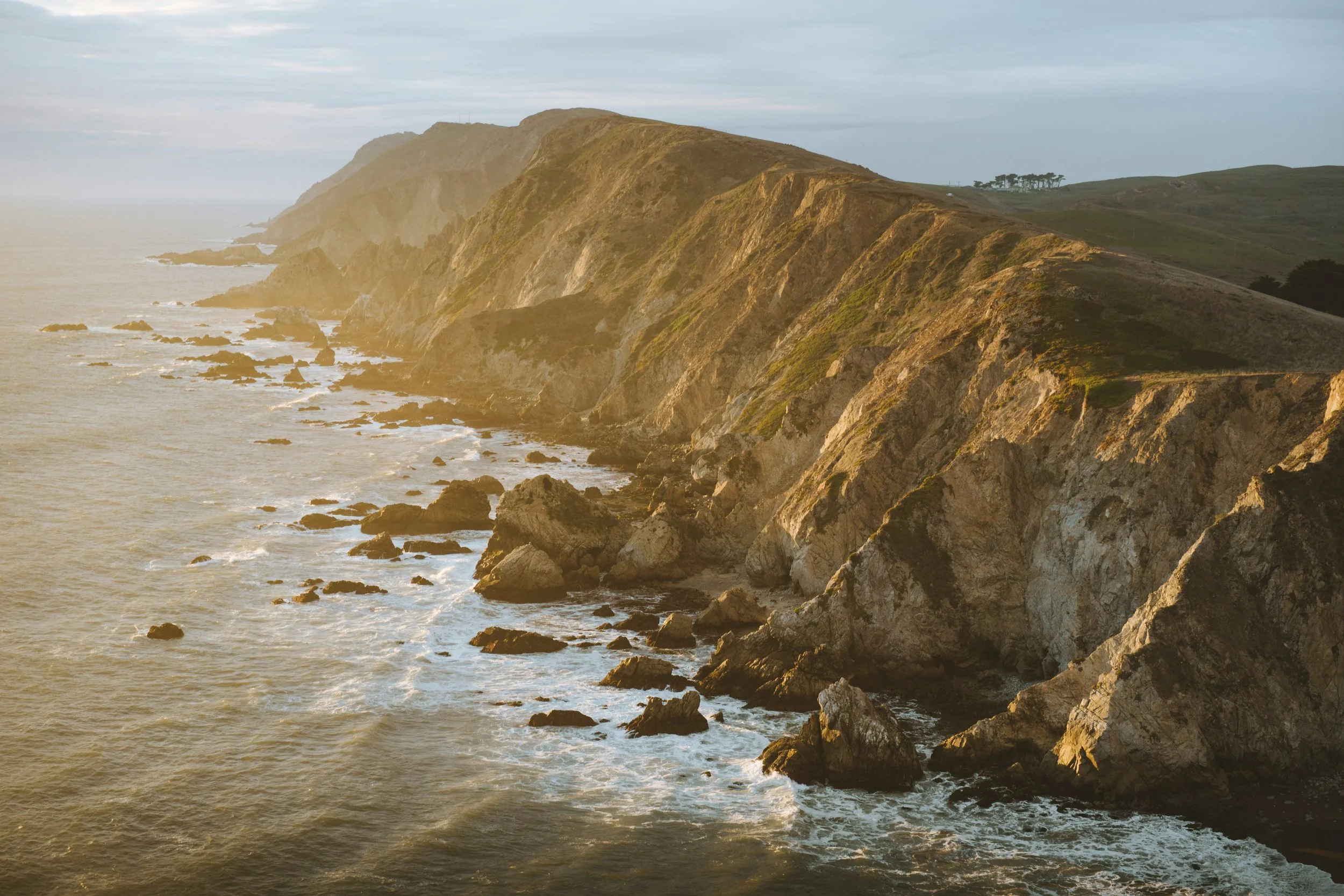 Cliffside overlooking the ocean during sunset with rocky shoreline and distant trees.
