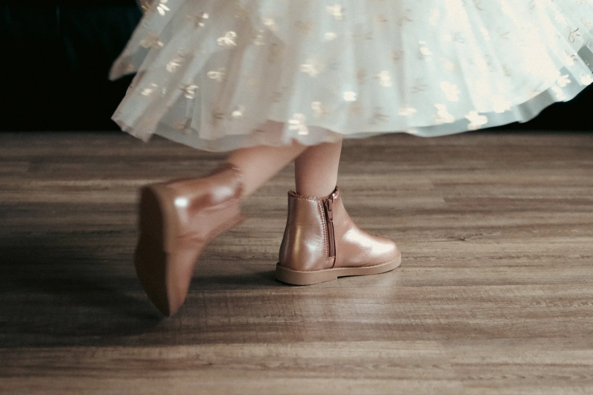 Close-up of a person's legs and feet, showing they are wearing shiny, light-colored ankle boots and a dress with a light, transparent fabric and metallic dot pattern, walking on a wooden floor.