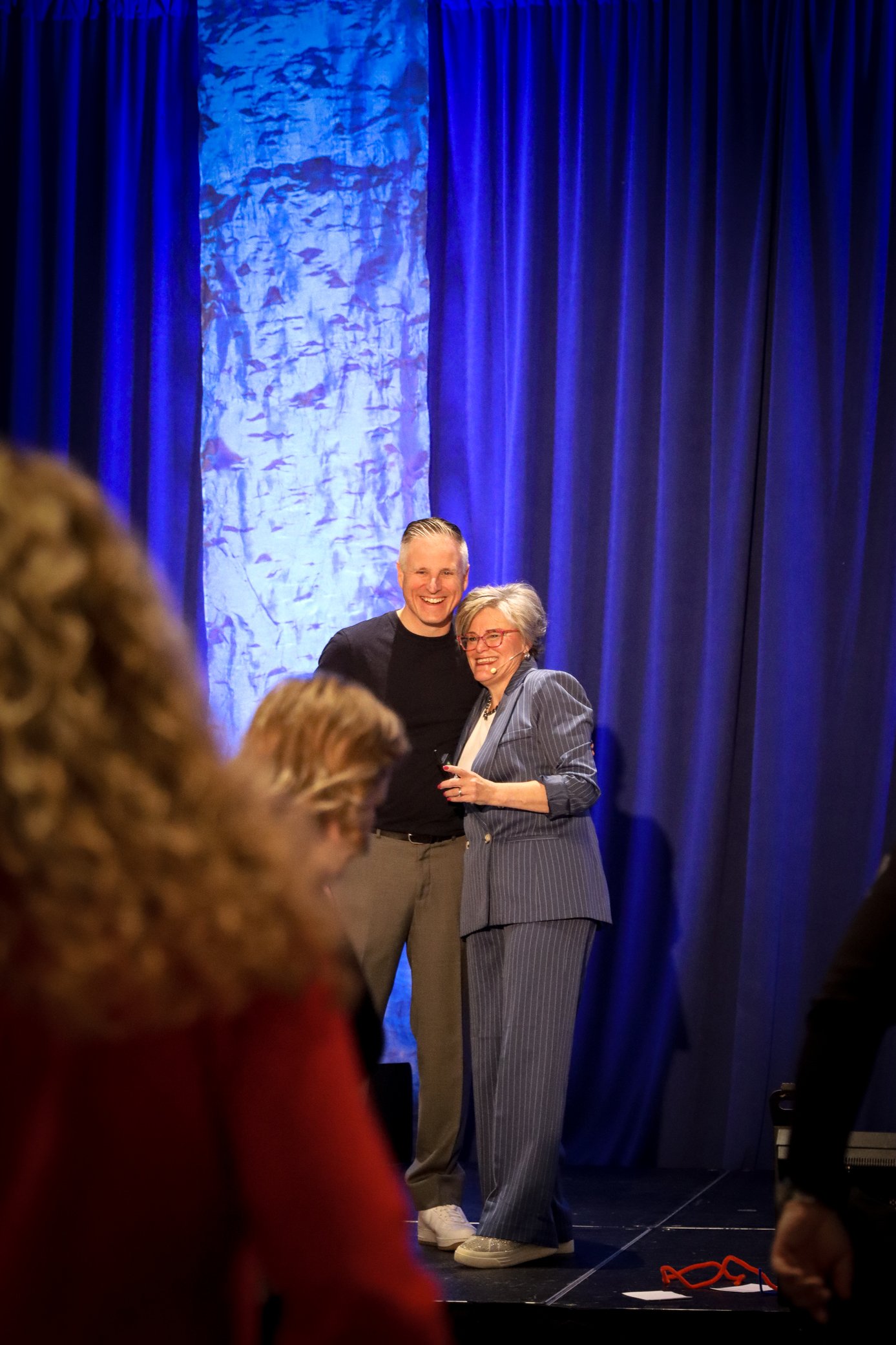 Two people, a man and a woman, standing on stage in front of blue curtains and a textured backdrop. They are smiling and posing for a photo. The woman is wearing a pinstripe suit and glasses. The man has short, slicked-back hair and is dressed in dar