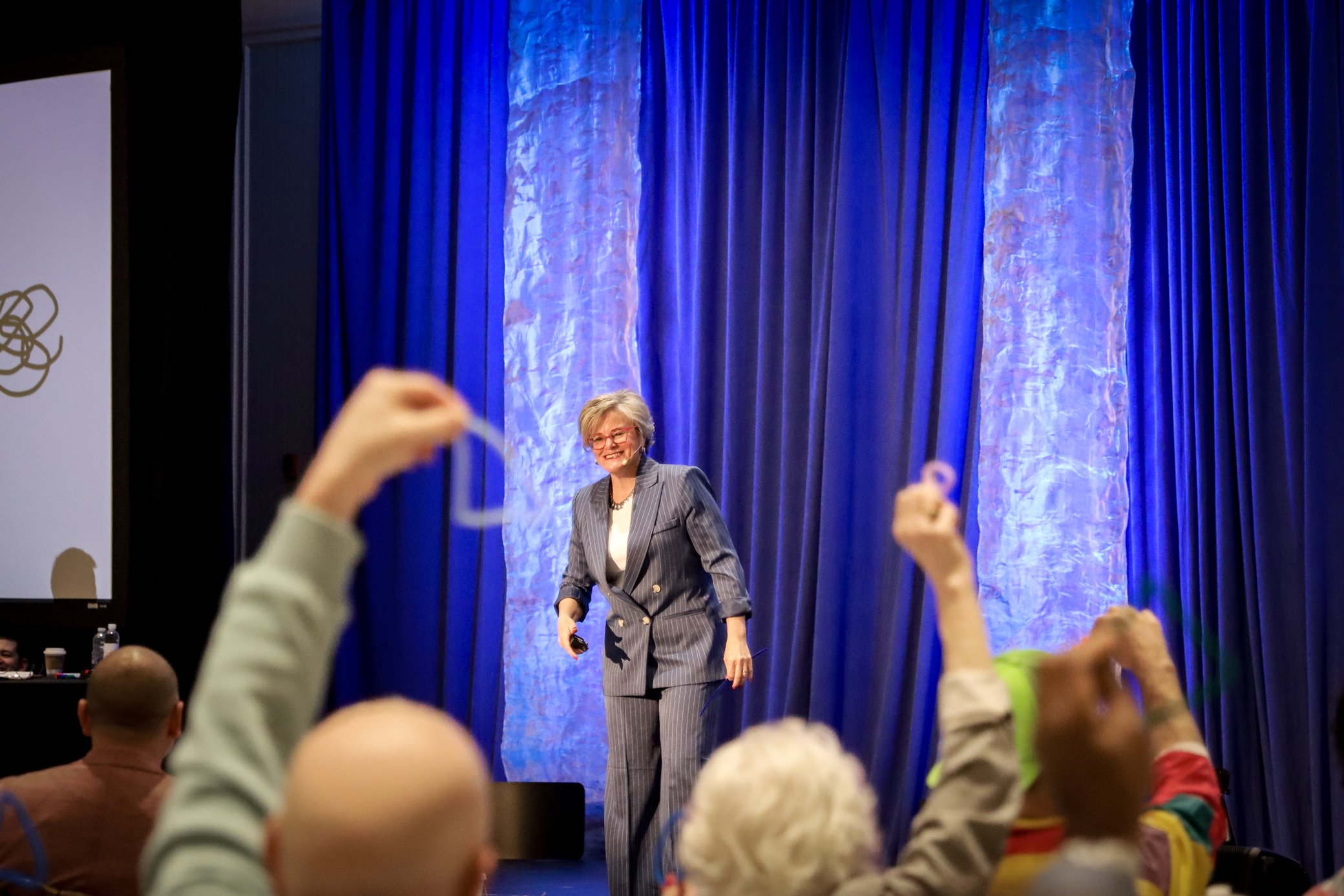 A woman in a striped suit smiling on a stage with blue curtains, with children raising their hands in the foreground.