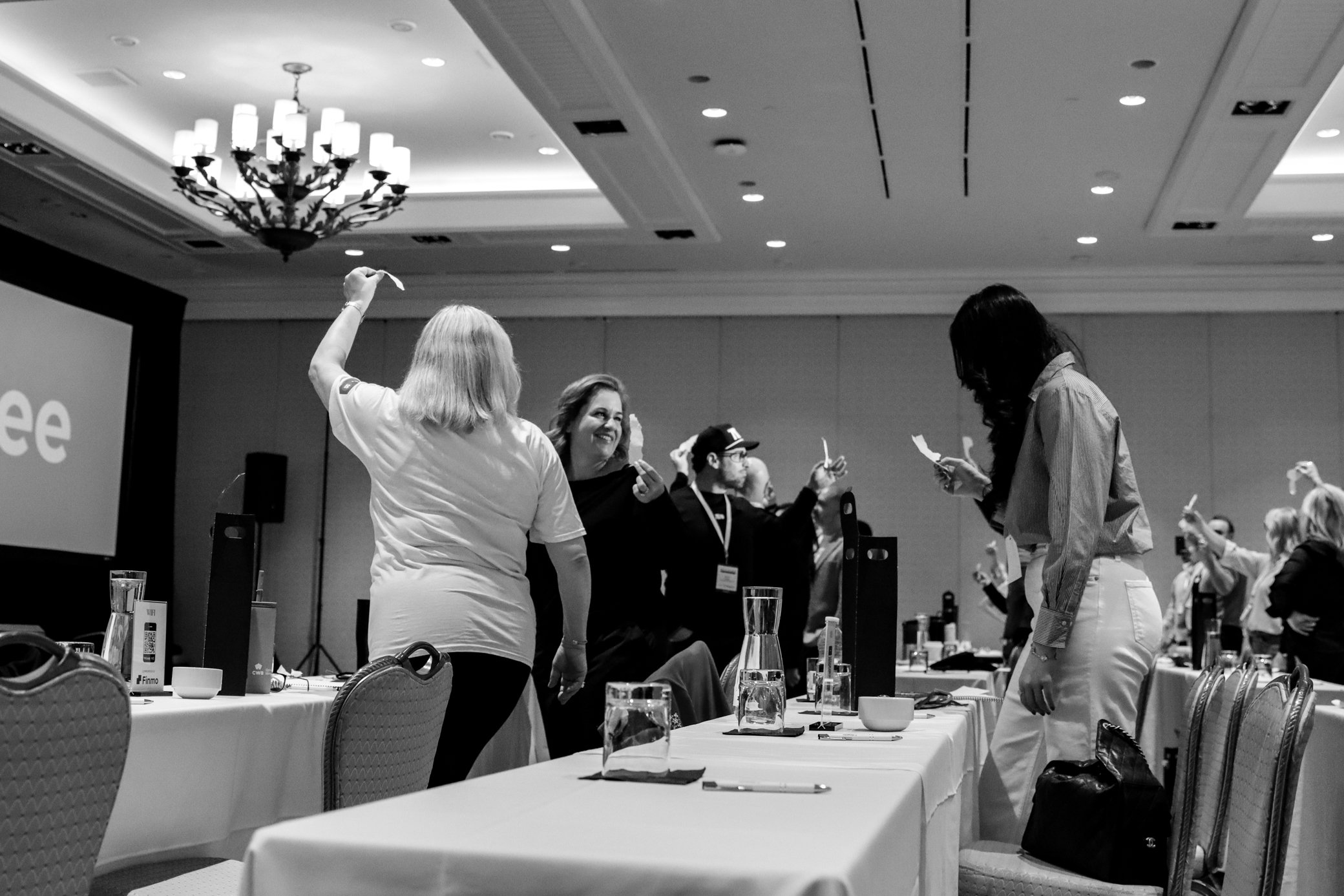 People standing and engaging with each other in a conference room with tables, chairs, and glasses of water on the tables.