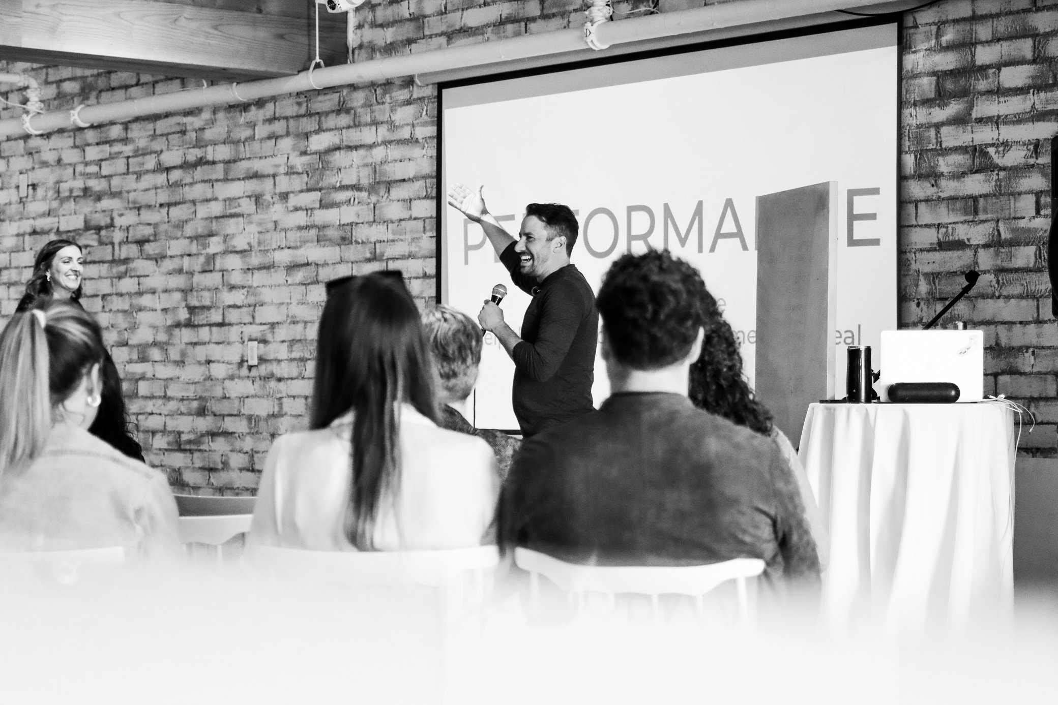 A man giving a presentation in front of a large screen with the word 'PERFORMANCE' on it, in a room with a brick wall and several seated audience members.