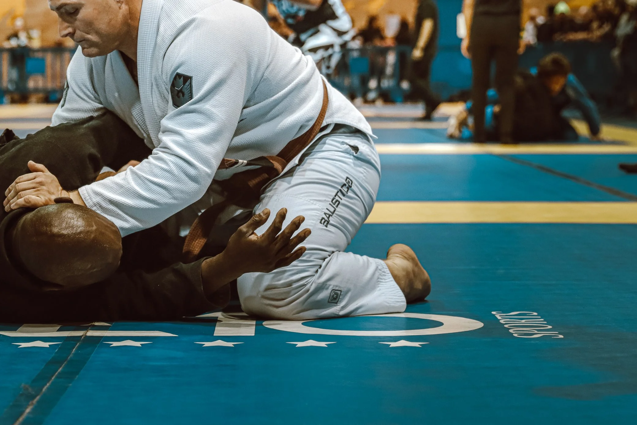 Brazilian Jiu-Jitsu practitioners wearing gis engaged in a match on a blue mat with white stars, with other competitors and spectators in the background.