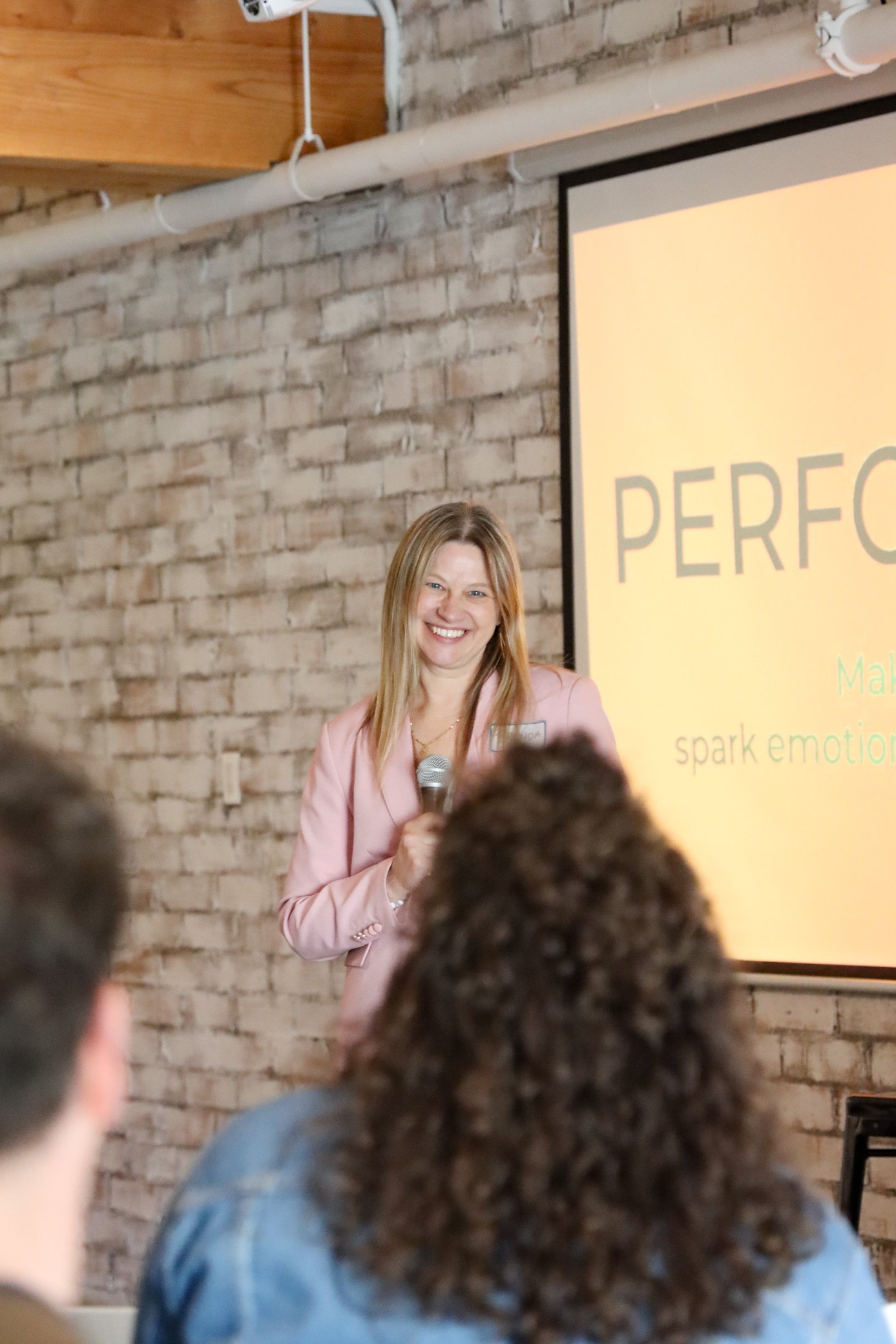 A woman in a pink blazer holding a microphone, smiling, standing in front of a projection screen with the partially visible word 'PERF' on a brick wall background during a presentation.