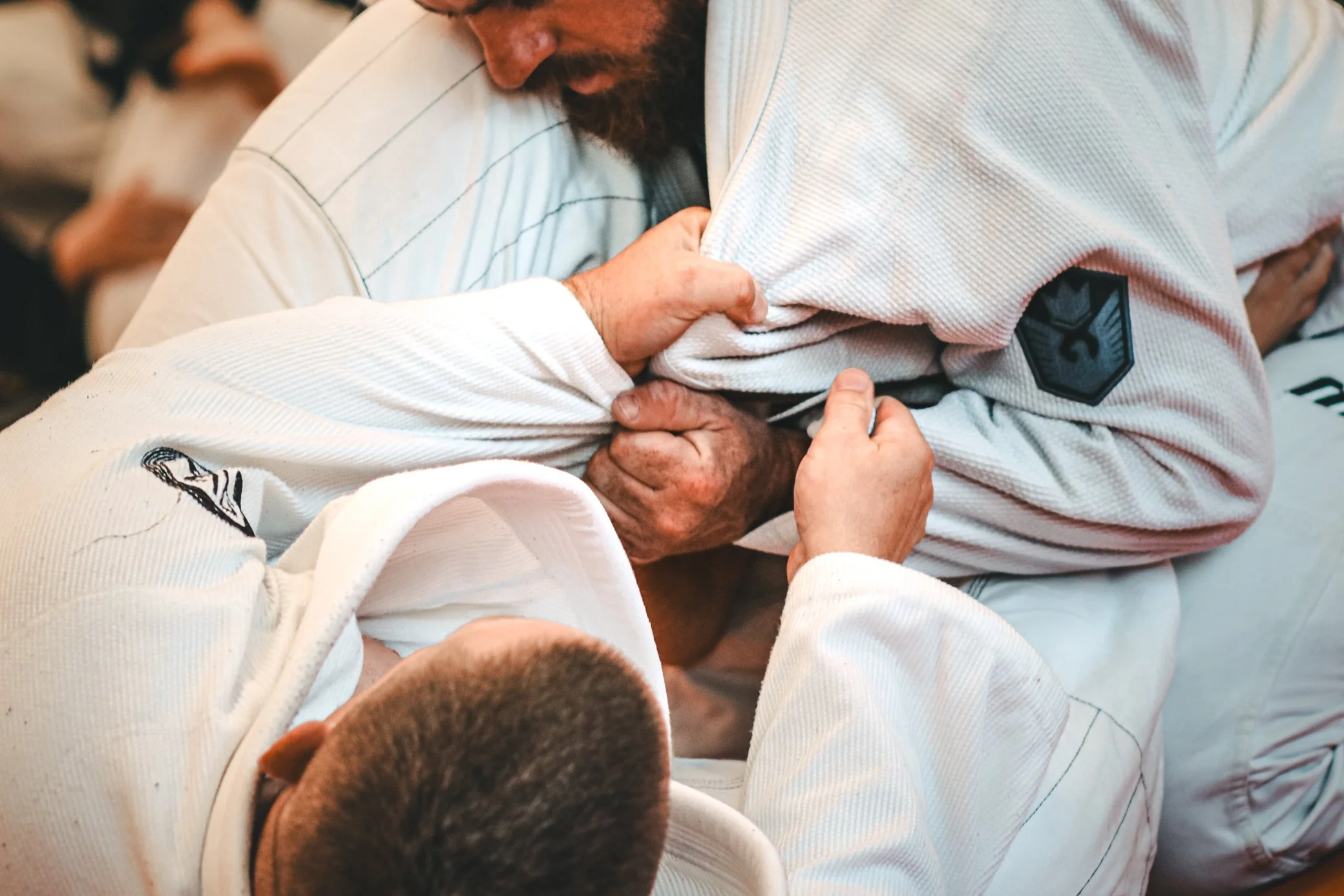 Two men engaged in a Brazilian Jiu-Jitsu match, one in a white gi with black patches and the other in a white gi with pinstripes, on a mat with a third participant nearby.
