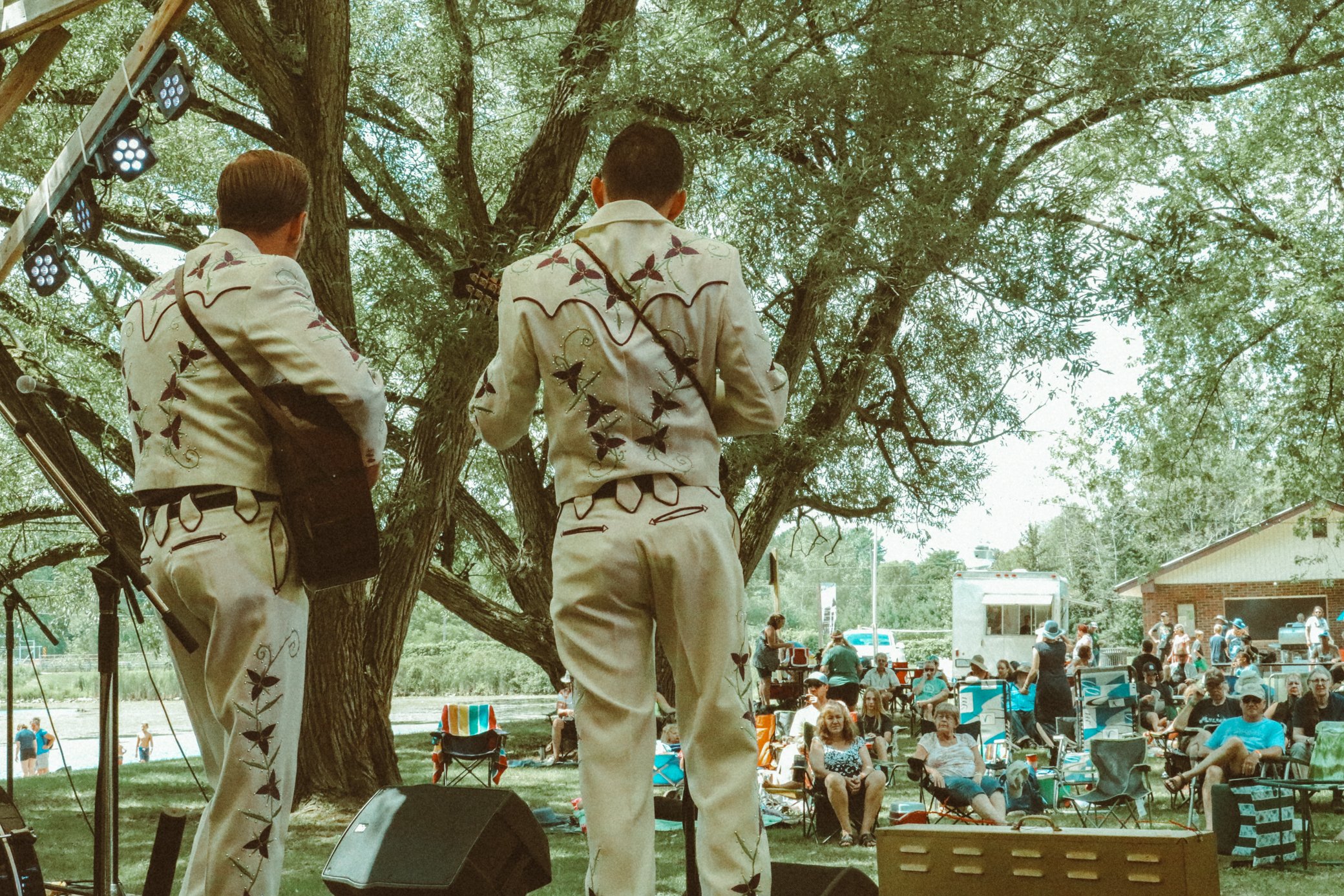 Two mariachi musicians performing outdoors on a stage under a large tree with an audience sitting and standing nearby on a sunny day.