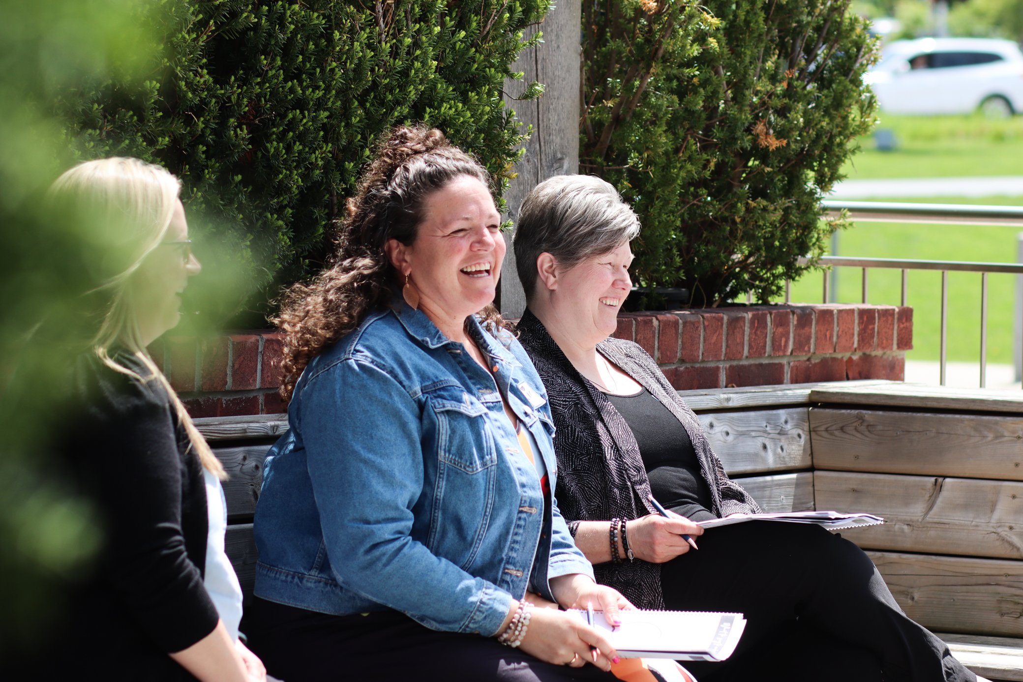Four women sitting on a wooden park bench near green bushes, some holding notebooks, and smiling outdoors on a sunny day.