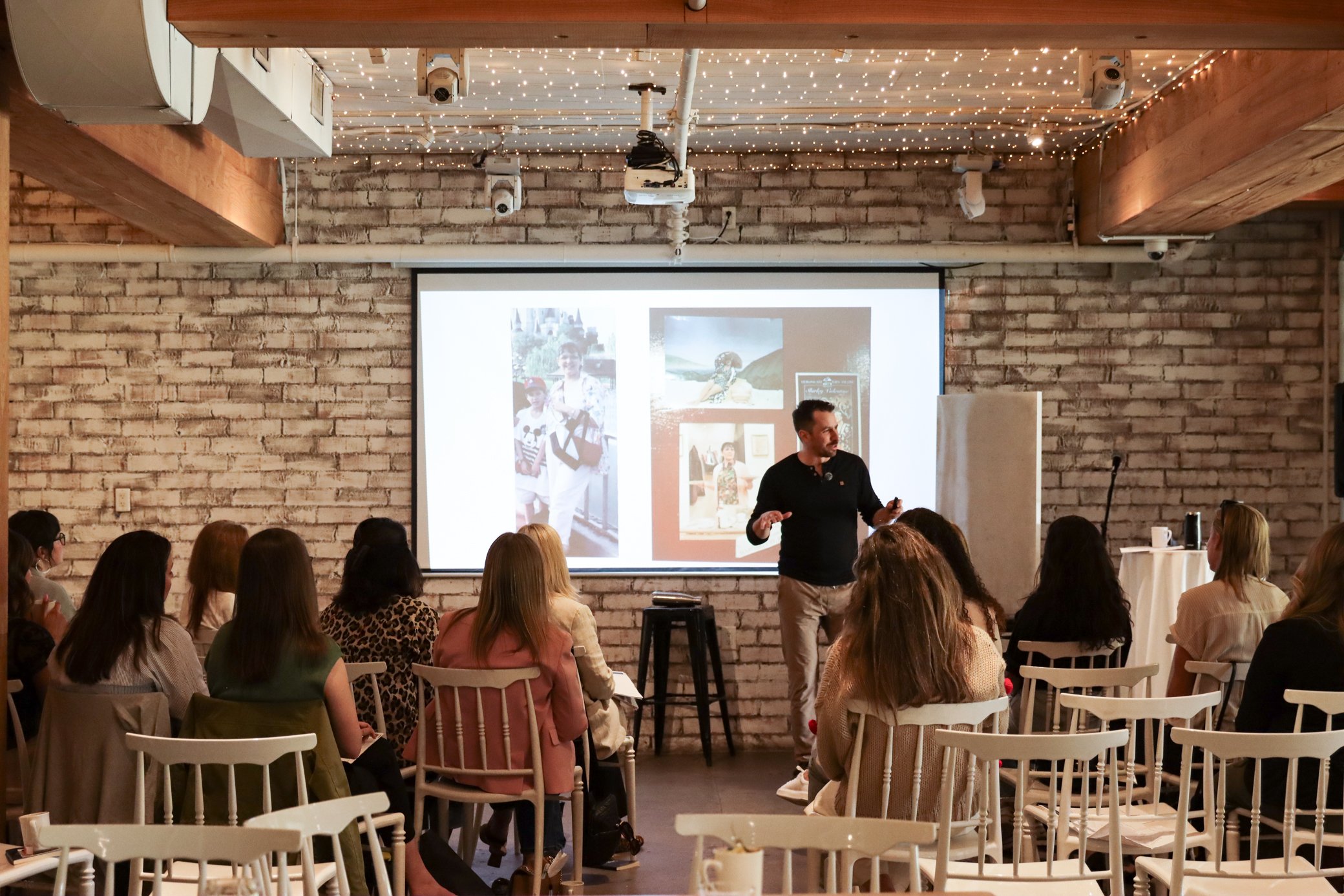 A man is giving a presentation in a room with exposed brick walls, decorated with string lights on the ceiling. He is standing in front of a large screen displaying photos of children. Attendees are seated facing him, listening attentively.