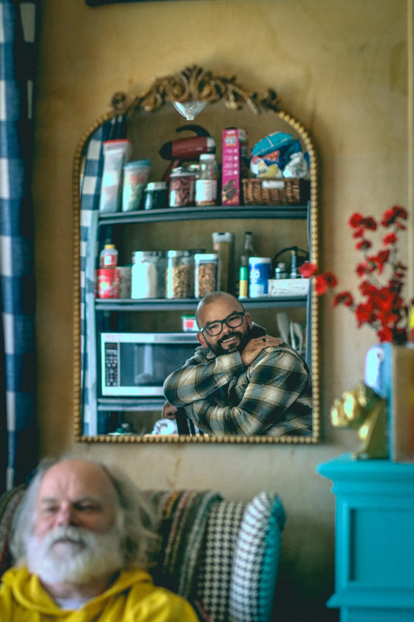 A man with glasses and a plaid shirt is smiling in a mirror reflection, while a bald man with a beard and a yellow hoodie is in the foreground, along with a partial view of an older man with a gray beard in the bottom left corner.