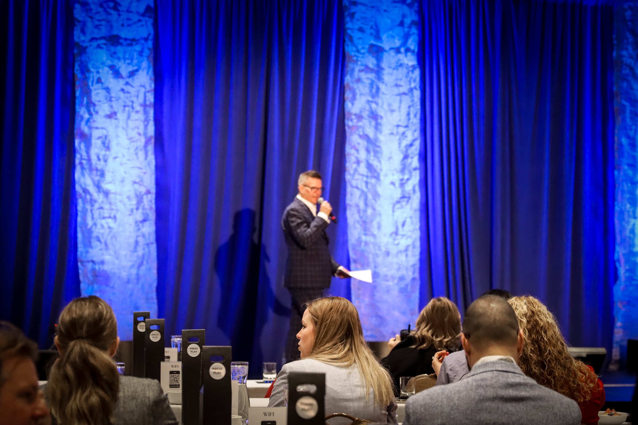 A man in a dark checkered suit and glasses speaking into a microphone on stage in front of blue curtains during an event. Audience members are seated at tables in front, with black signs indicating Wi-Fi, press, and play options.