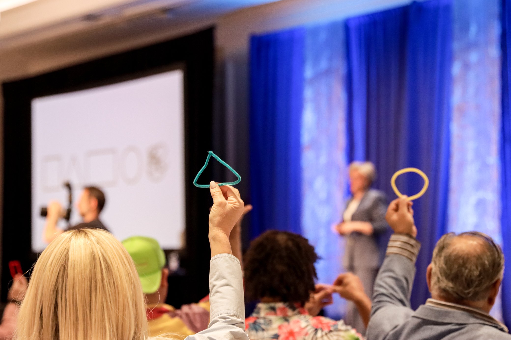 Audience at a presentation or conference raising their hands, some holding blue and yellow circular objects, with a speaker on stage in the background.
