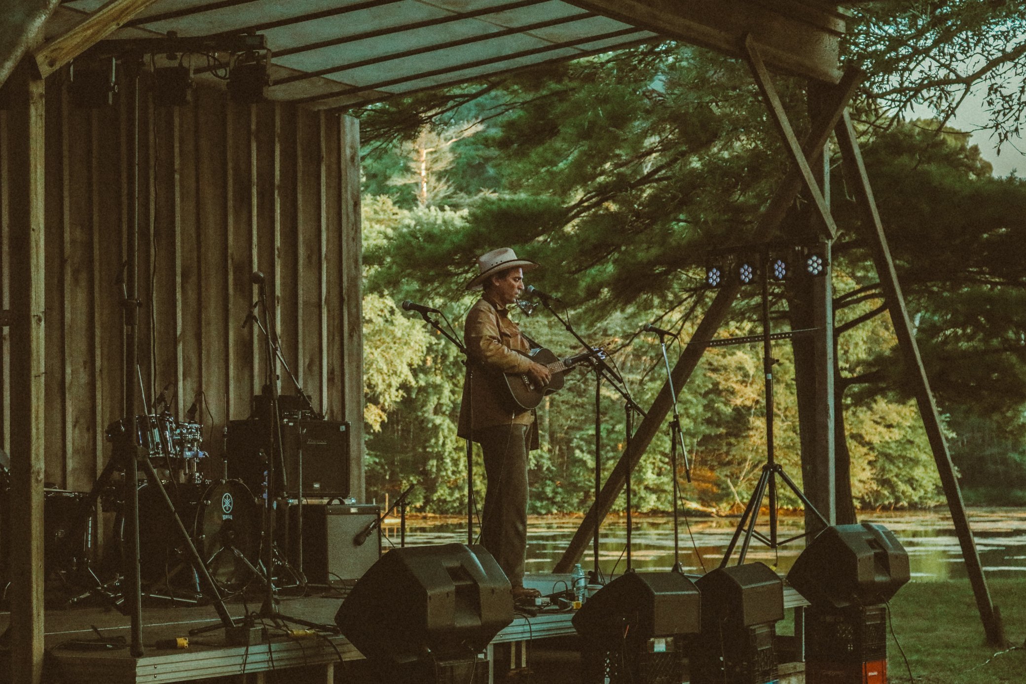 A musician performing on an outdoor stage surrounded by trees, with musical equipment and stage lights visible.
