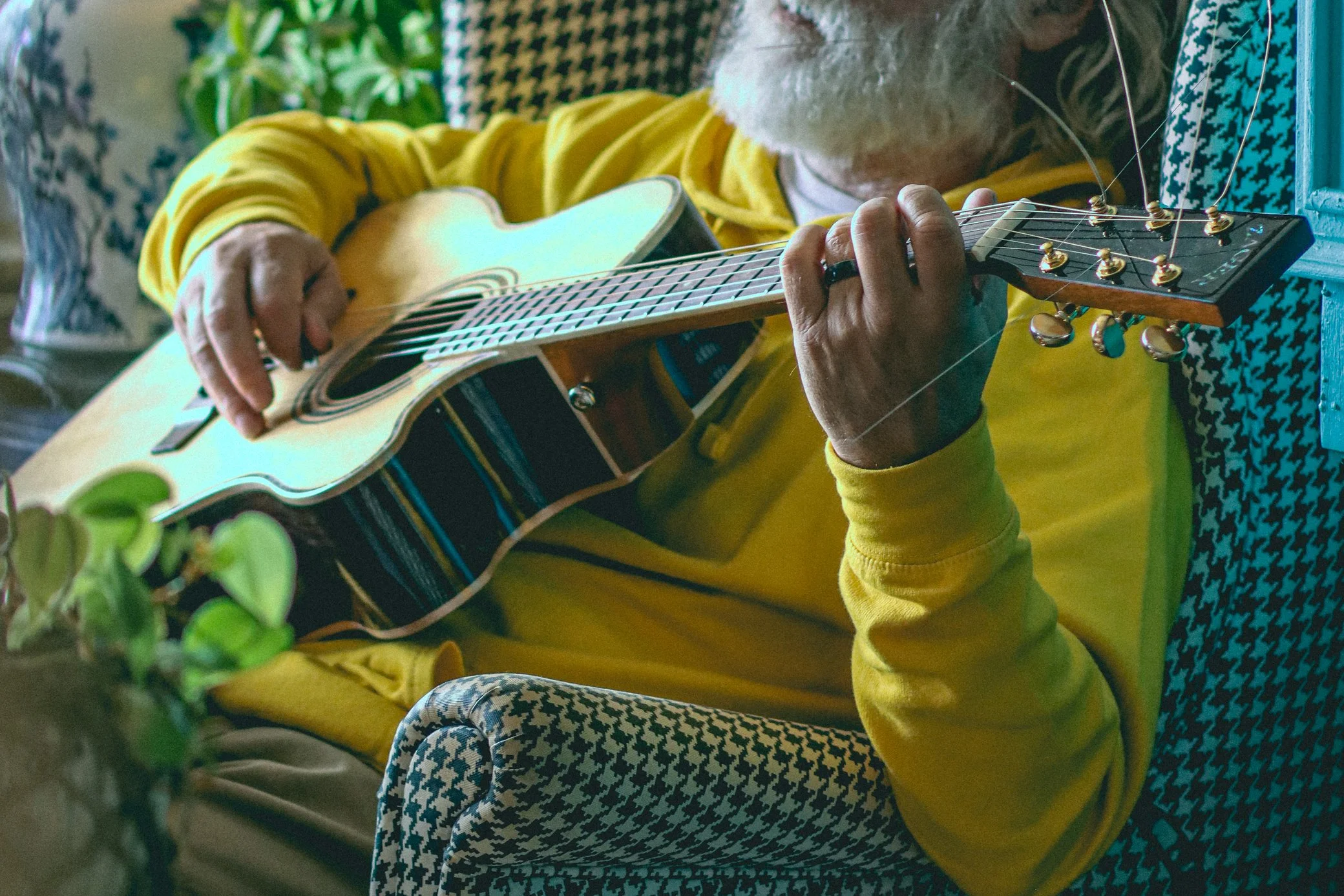 An elderly man with a gray beard and glasses playing an acoustic guitar while sitting on a chair. He is wearing a yellow sweater and patterned pants, and is positioned near a blue wall with a houndstooth cushion and some green plants nearby.