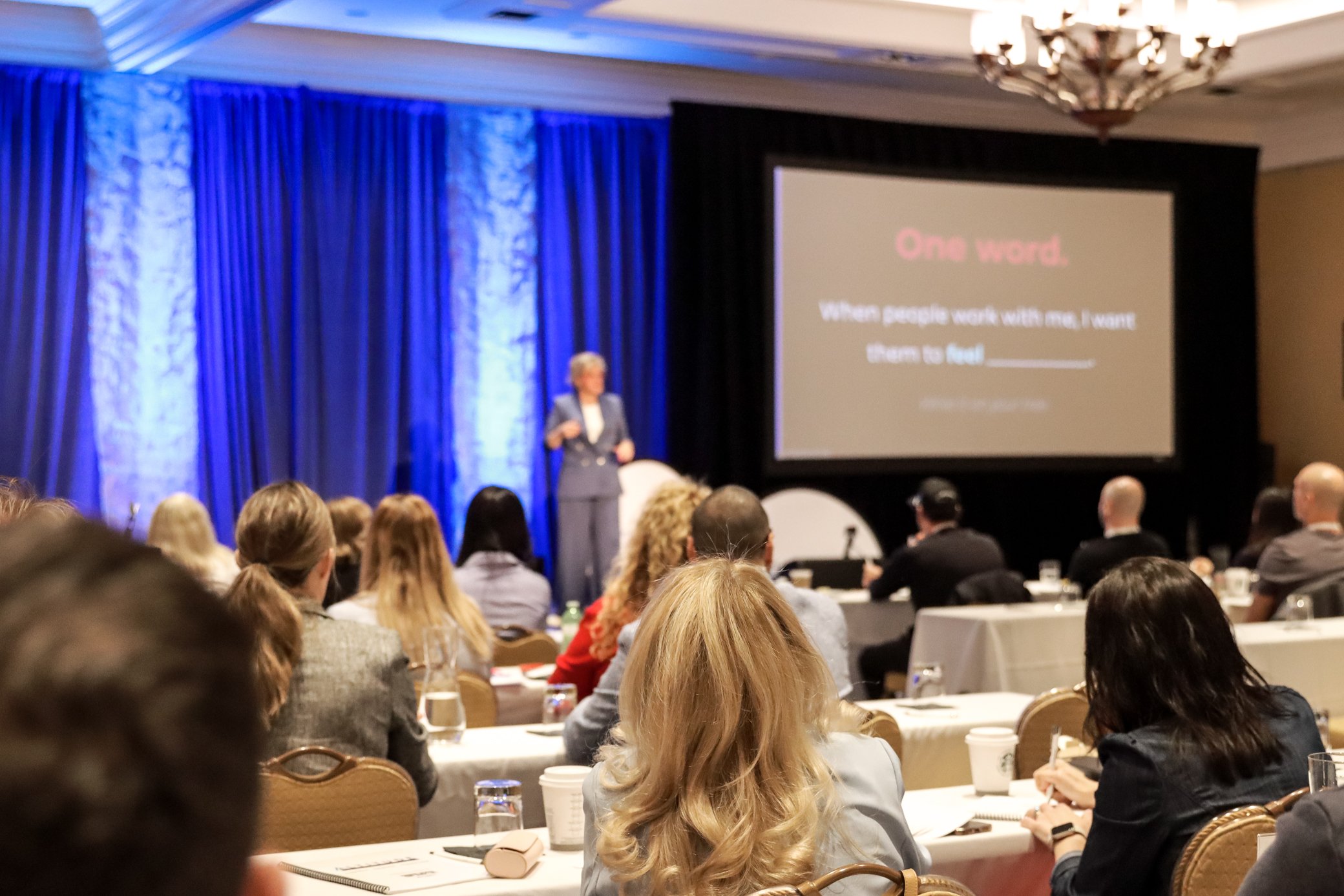 A woman standing and speaking on stage during a conference or seminar, with a large screen displaying presentation text that says "One word" and a quote about the importance of one word. The audience is seated at tables, listening and taking notes in