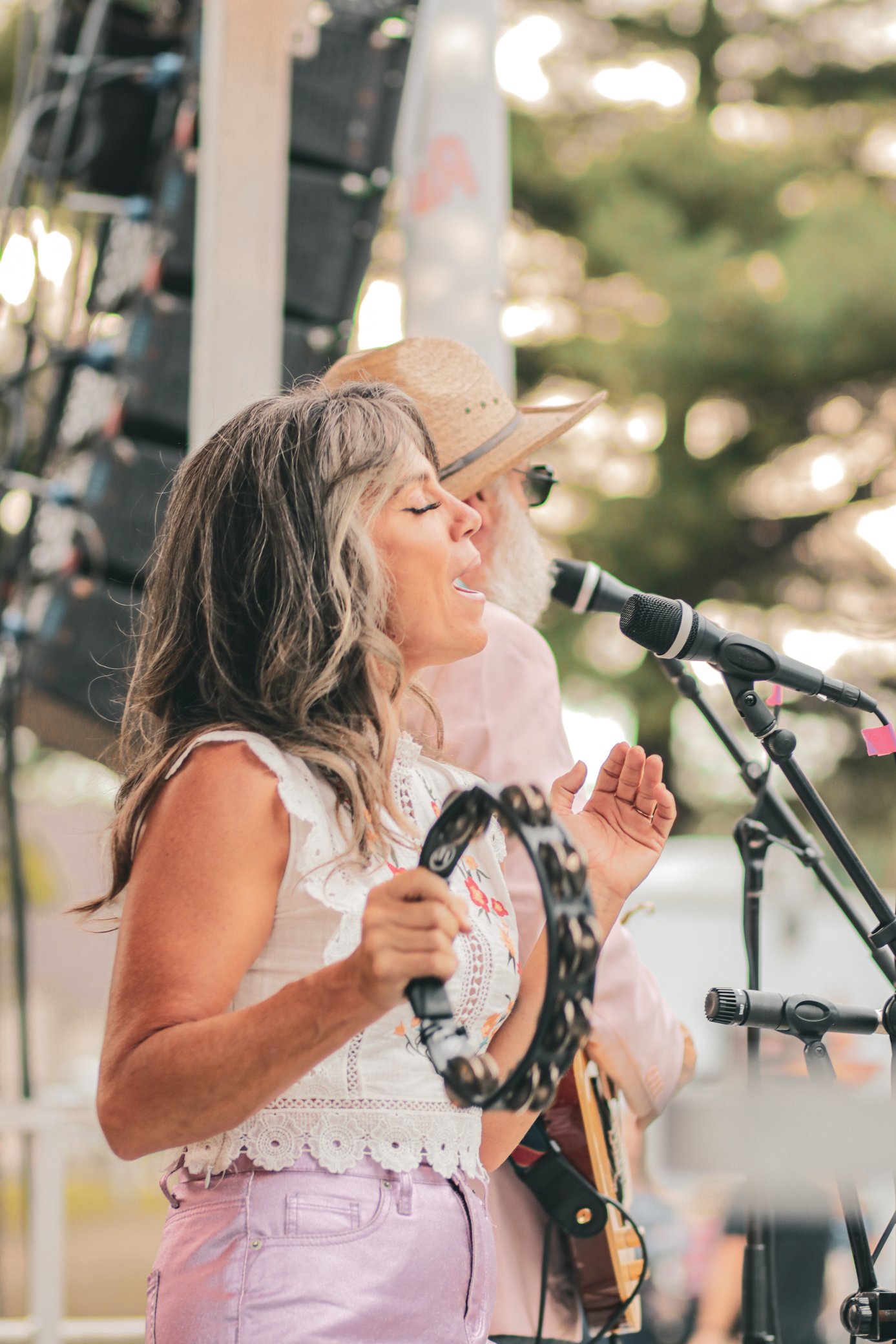 Two women, one with gray hair and the other with long, dark hair wearing a straw hat, are singing into microphones at an outdoor event surrounded by trees.
